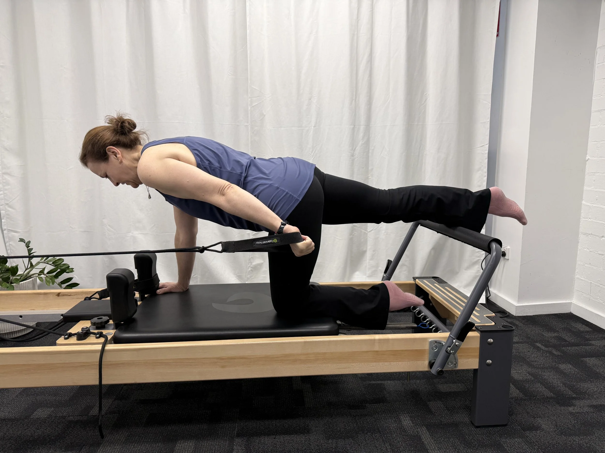 A woman with brown hair in a bun, wearing a blue sleeveless shirt and black leggings, performing a yoga or exercise pose on a reformer machine indoors with a white curtain backdrop and some plants in the corner.
