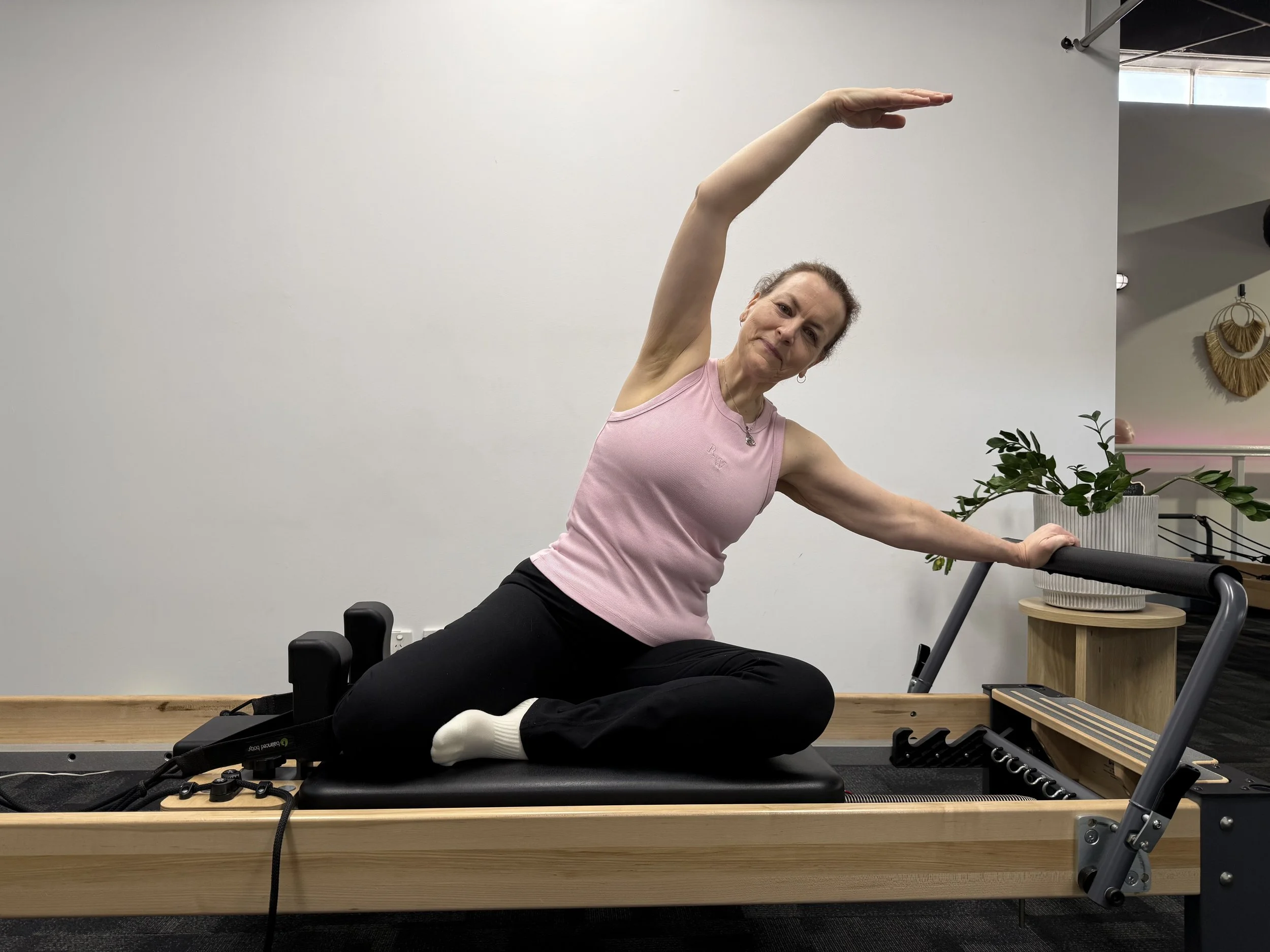 A woman in a pink sleeveless top and black pants practicing Pilates on a reformer machine in a studio with minimal decor, including a plant and decorative wall hanging.