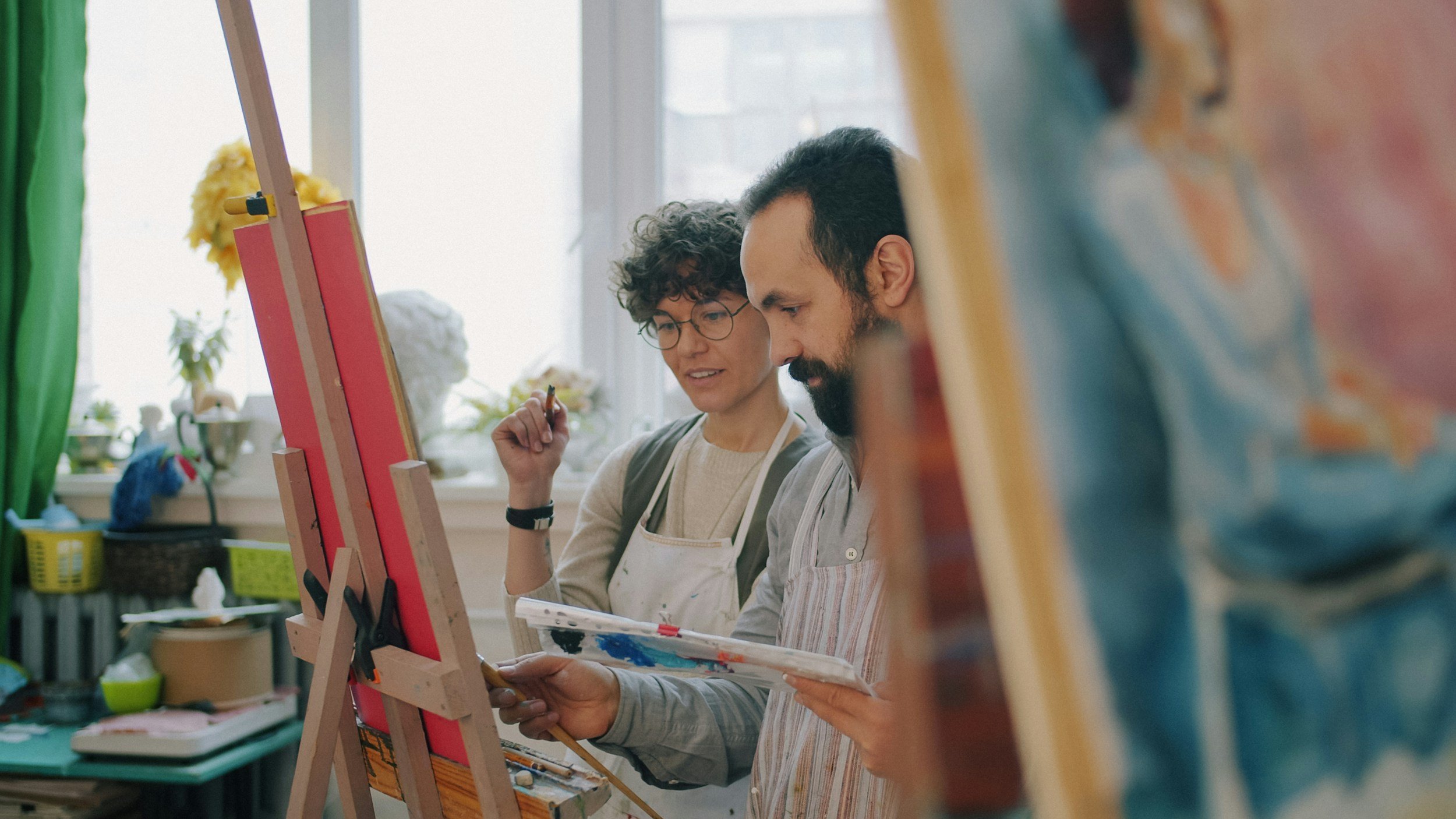 Two people painting in an art studio, standing close to each other and looking at their paintings on easels.