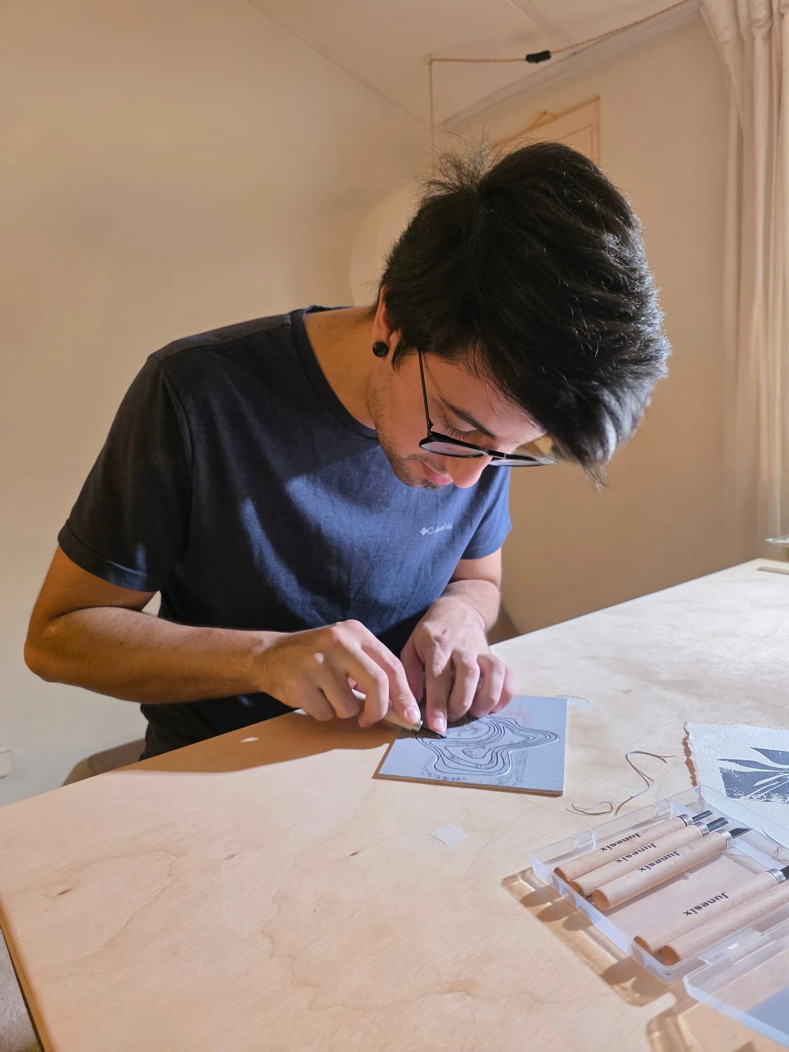 The artist, a man with black hair, glasses, and a grey shirt is carving a piece of lino while sitting at a wooden table. There are beige carving tools in a plastic case on the table, and the background includes a white wall and curtains.