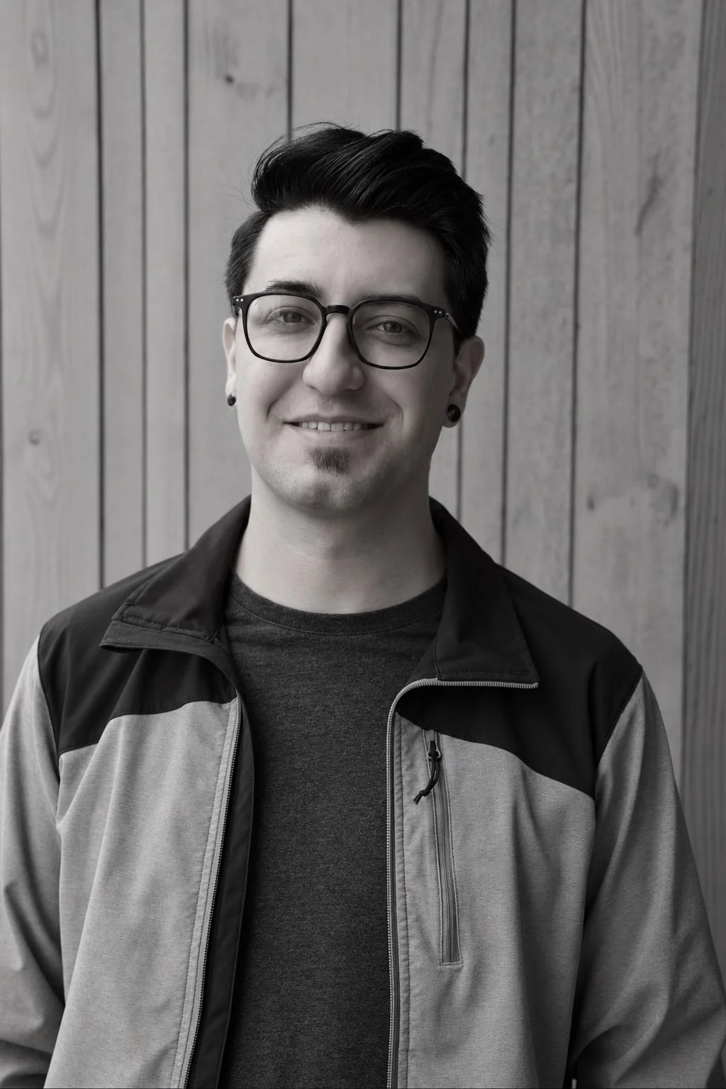 Portrait of the artist, Avery, a young man with glasses, black ear gauge earrings, and styled hair, smiling, standing in front of a wooden wall.