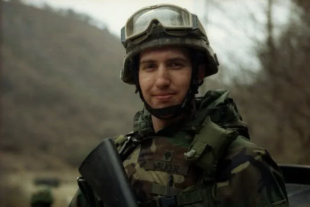 A soldier wearing camouflage uniform, helmet, and goggles, standing outdoors in a wooded area.