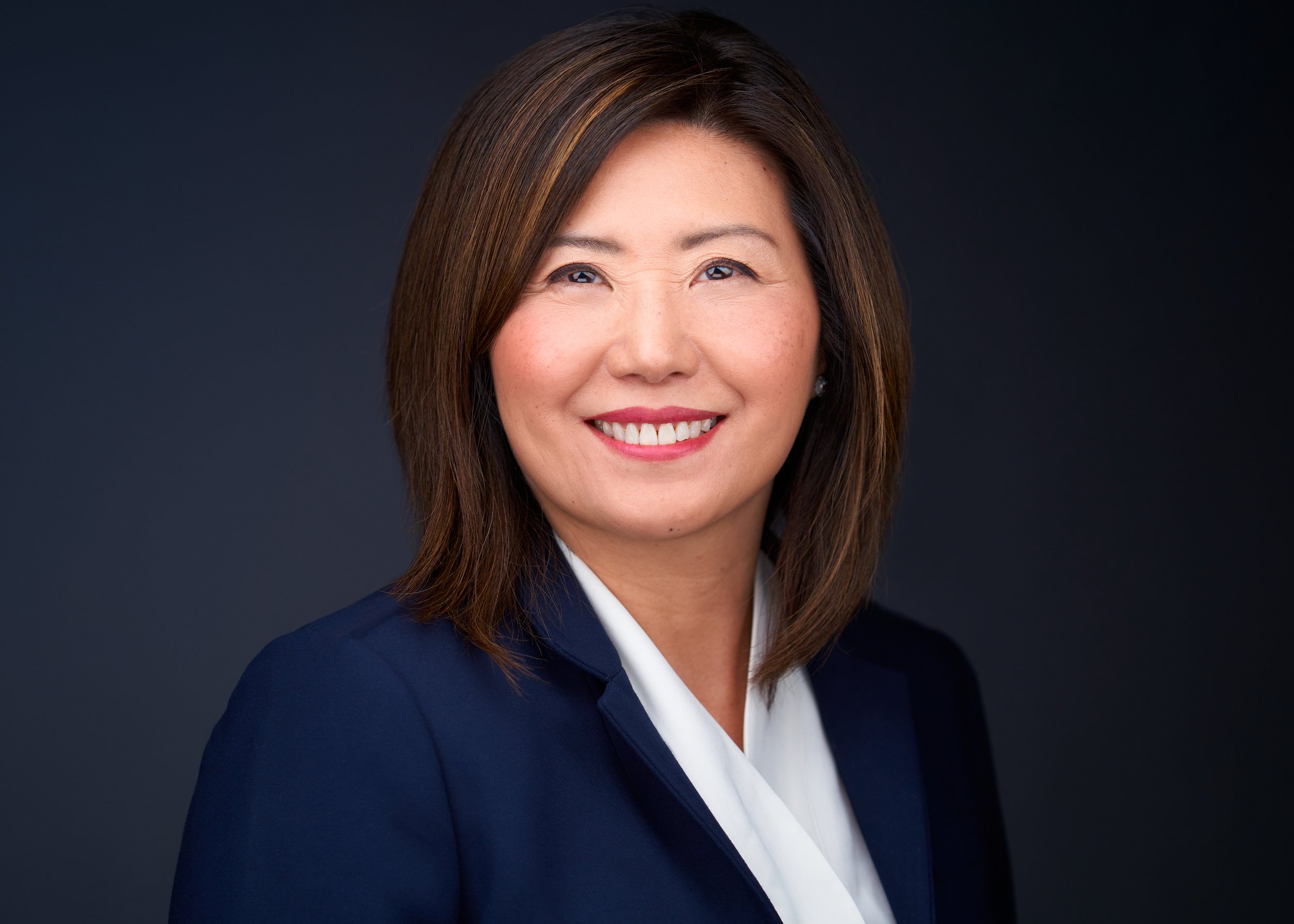 Professional woman with shoulder-length brown hair, wearing a navy blazer and white shirt, smiling against a dark background.