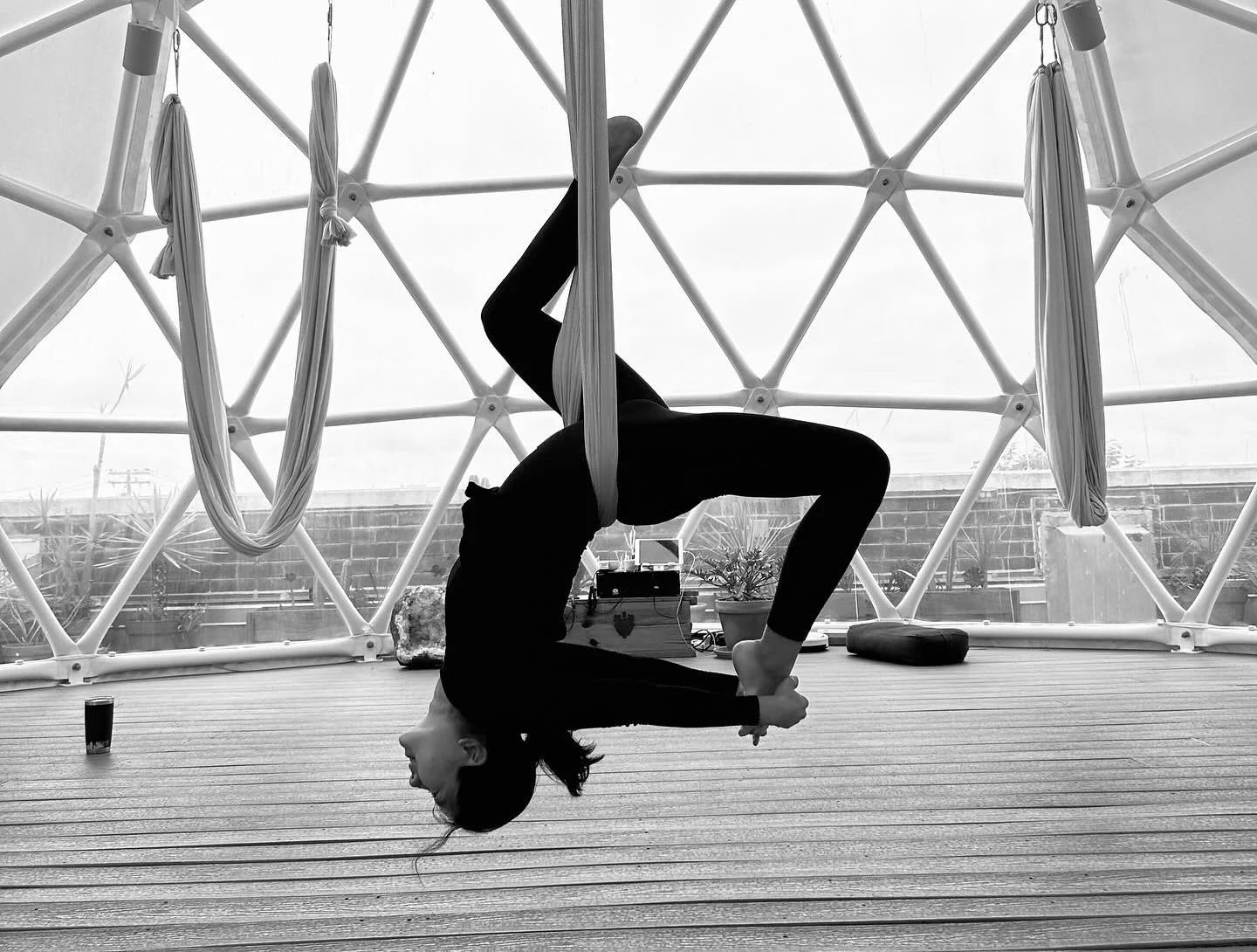 A person in black yoga attire performs an aerial yoga pose in a geodesic dome with fabric swings hanging from the structure, outdoors on a deck with potted plants and a view of a cloudy sky.