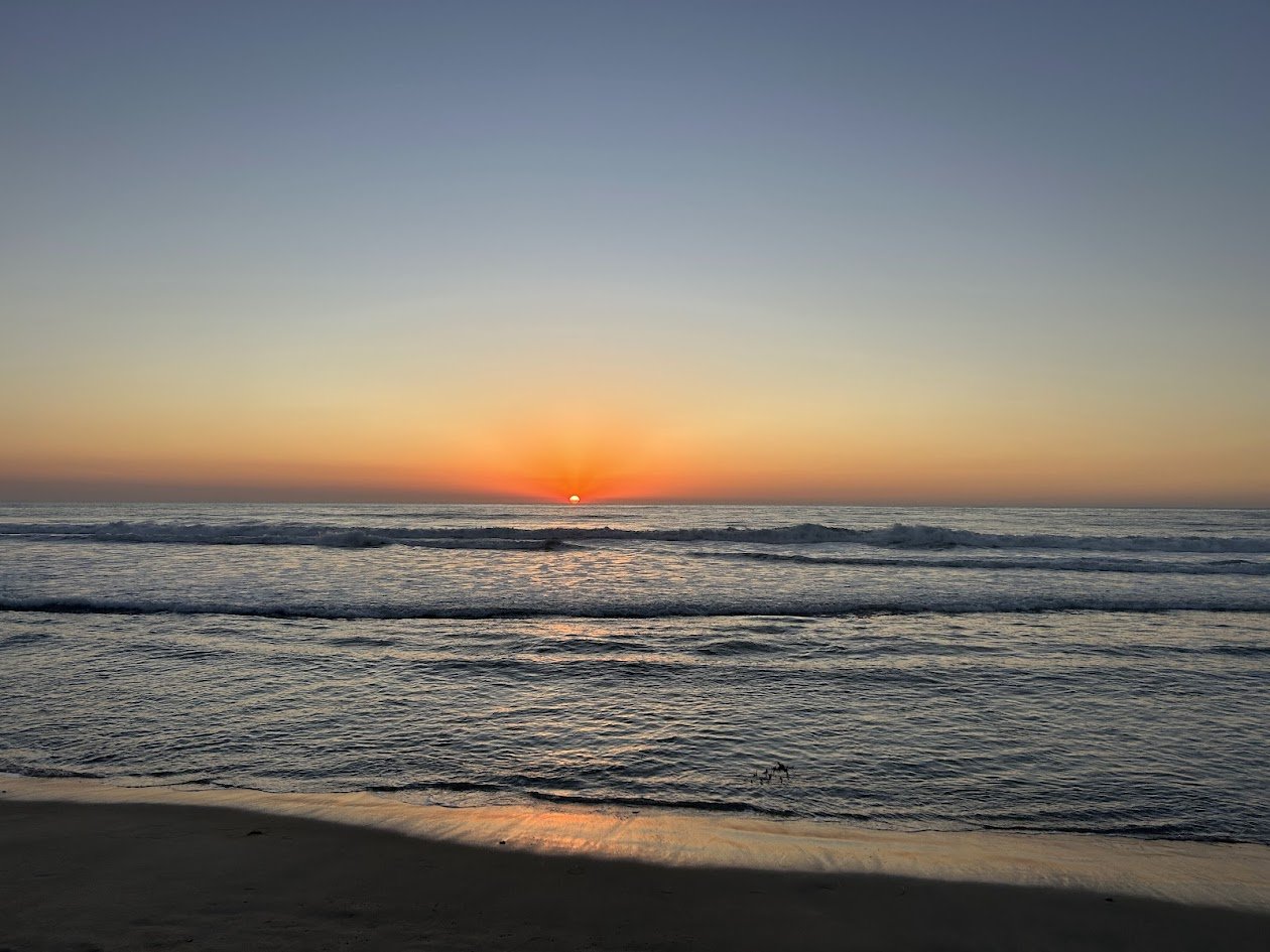 Sunset over the ocean with gentle waves and a sandy beach in the foreground.