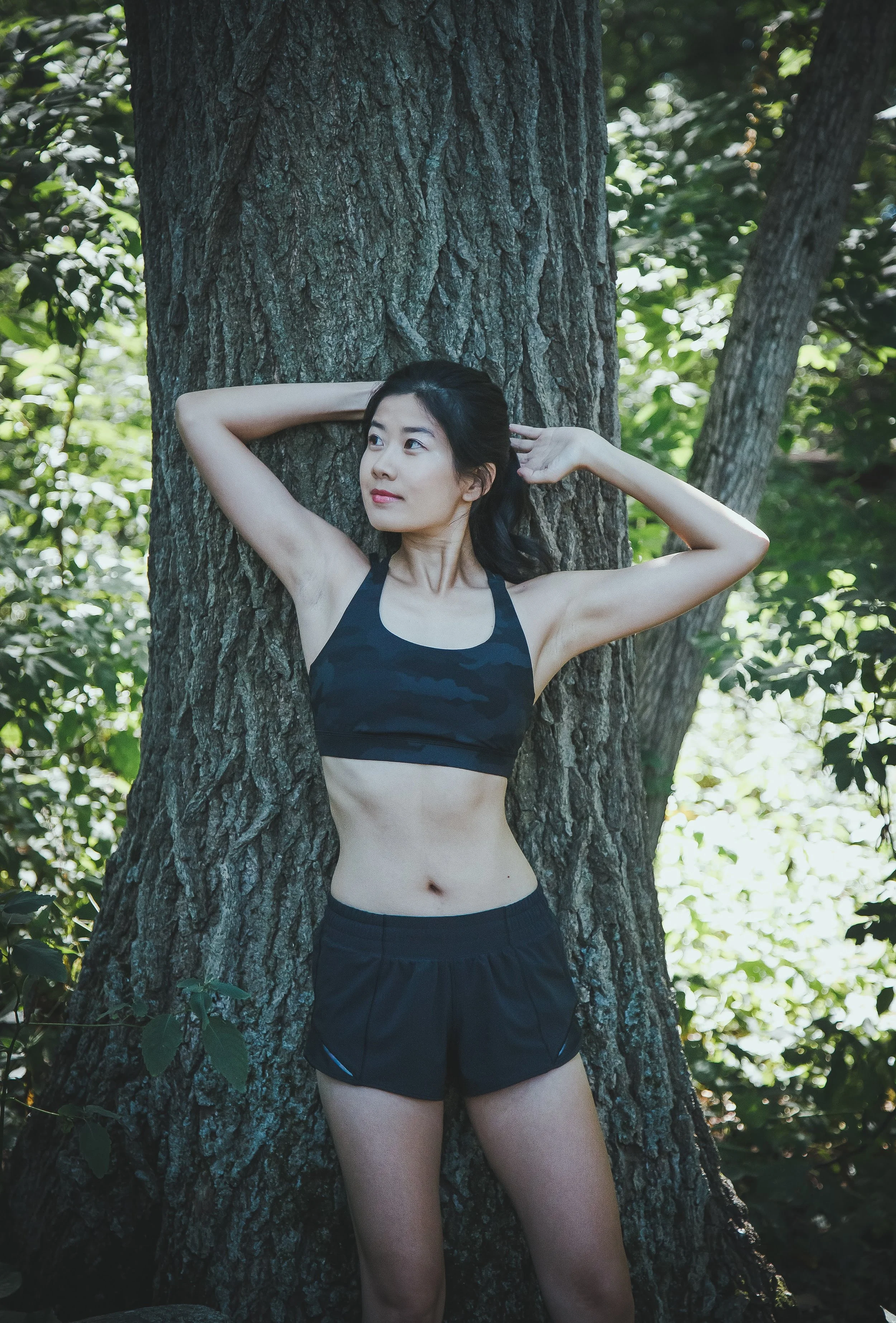 A woman in athletic clothing leaning against a large tree trunk outdoors, with dense green foliage in the background.