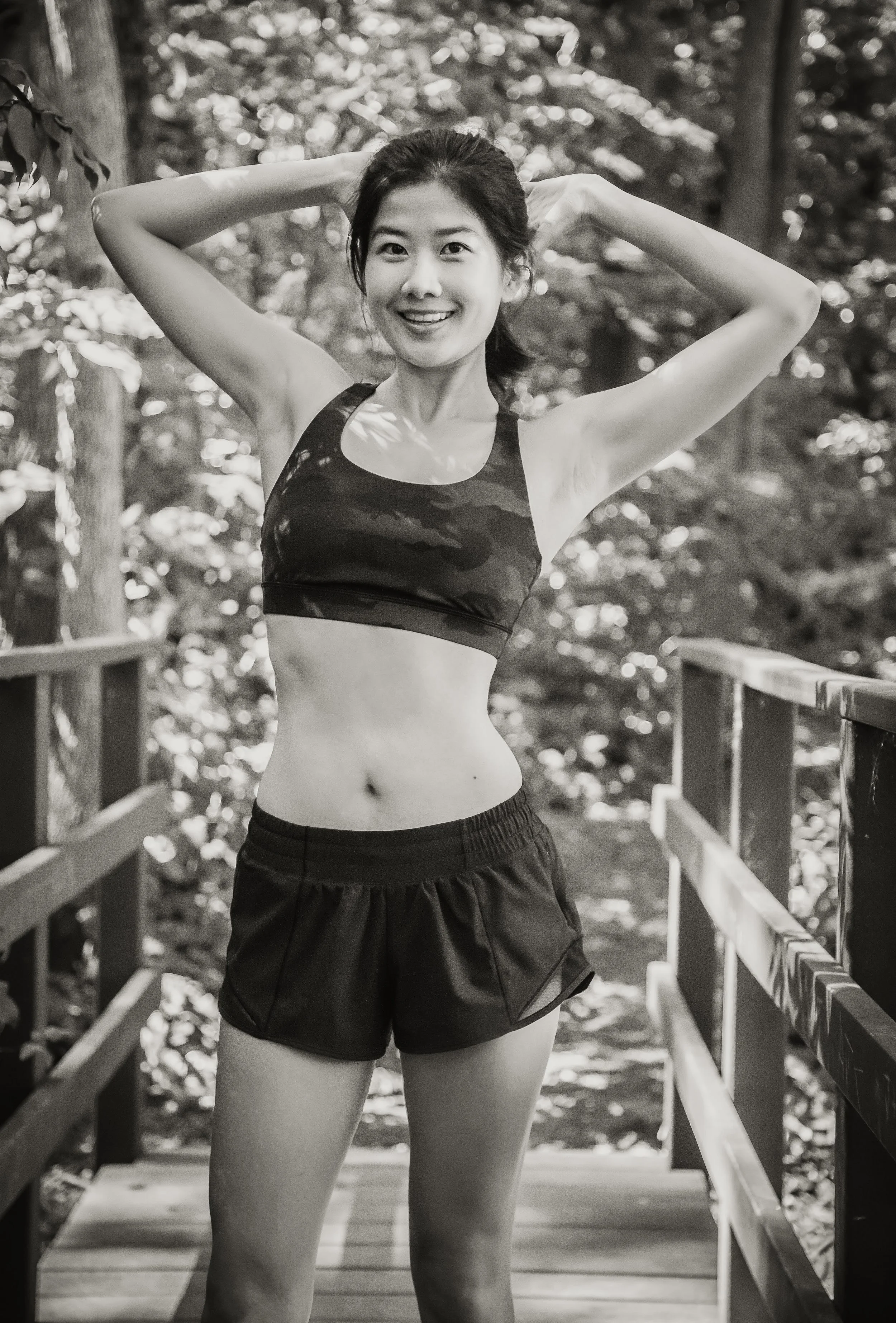 A young woman is standing outdoors on a wooden bridge, smiling, with her arms raised behind her head, wearing a sports bra and shorts, in a shaded forest setting.