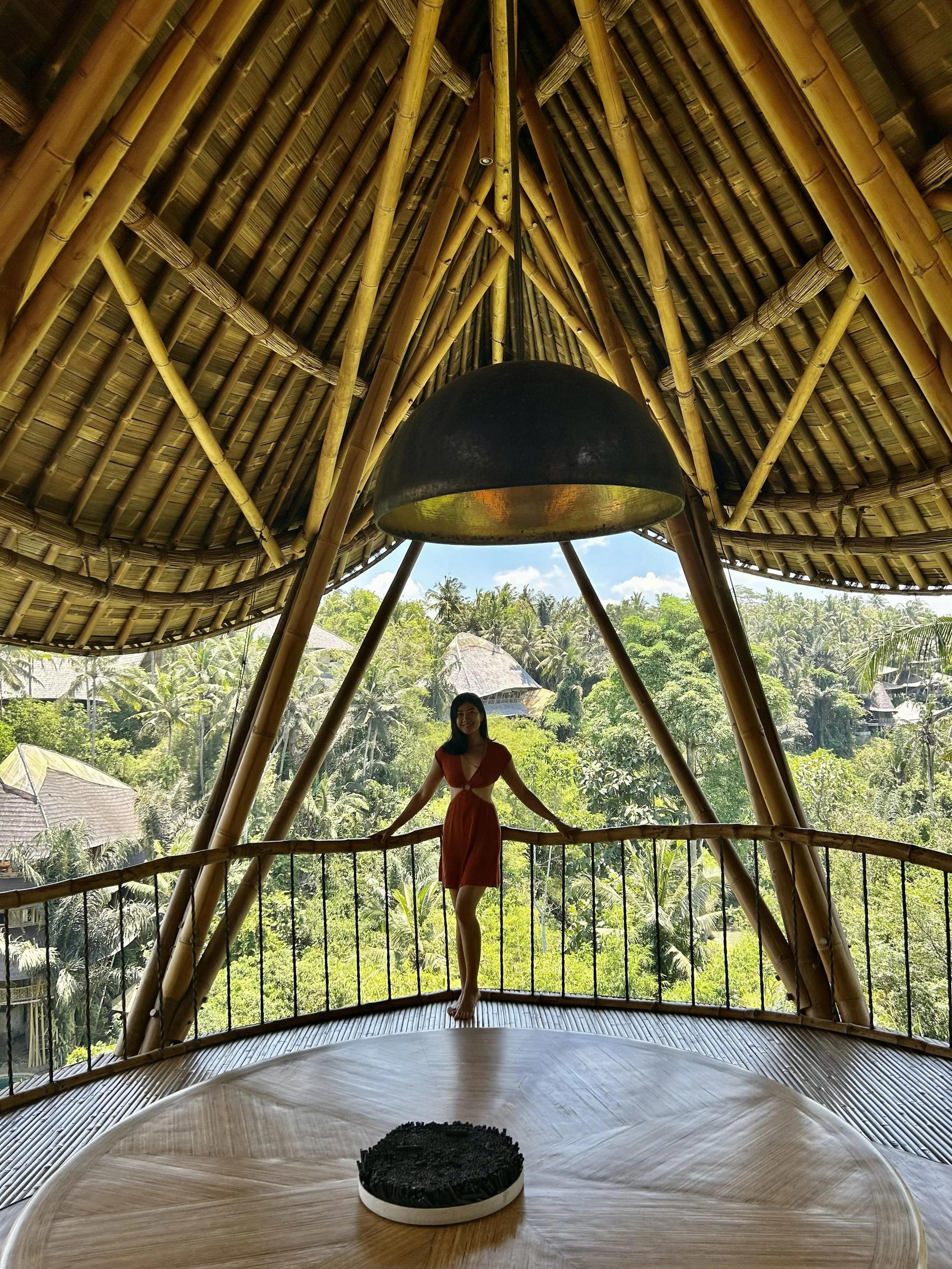 A woman in a red dress standing on a bamboo balcony with a tropical jungle view behind her. The interior features a bamboo ceiling and a large black hanging light fixture.