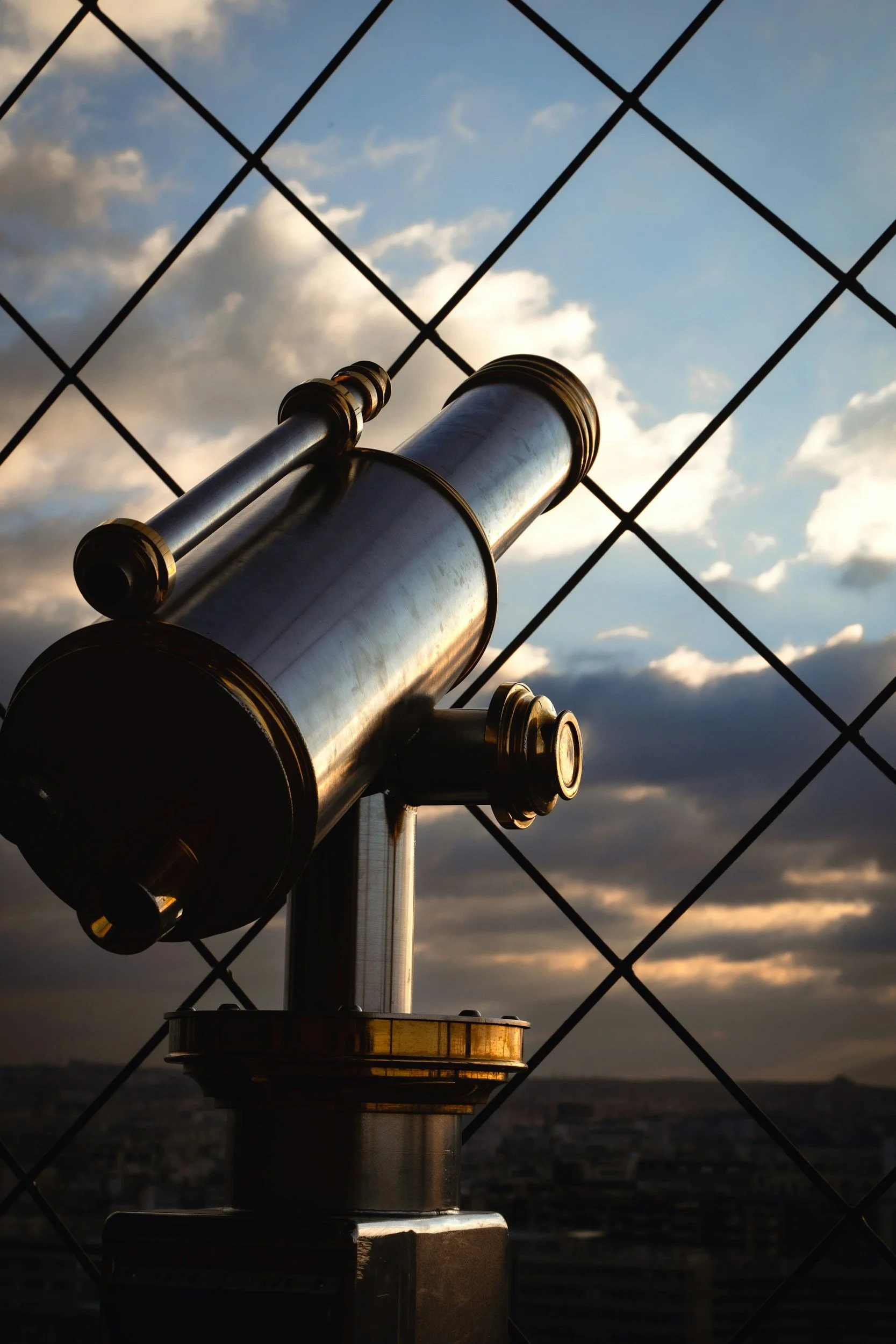 A telescope mounted on a rooftop, viewed from below, with a cityscape and a partly cloudy sky at sunset in the background.