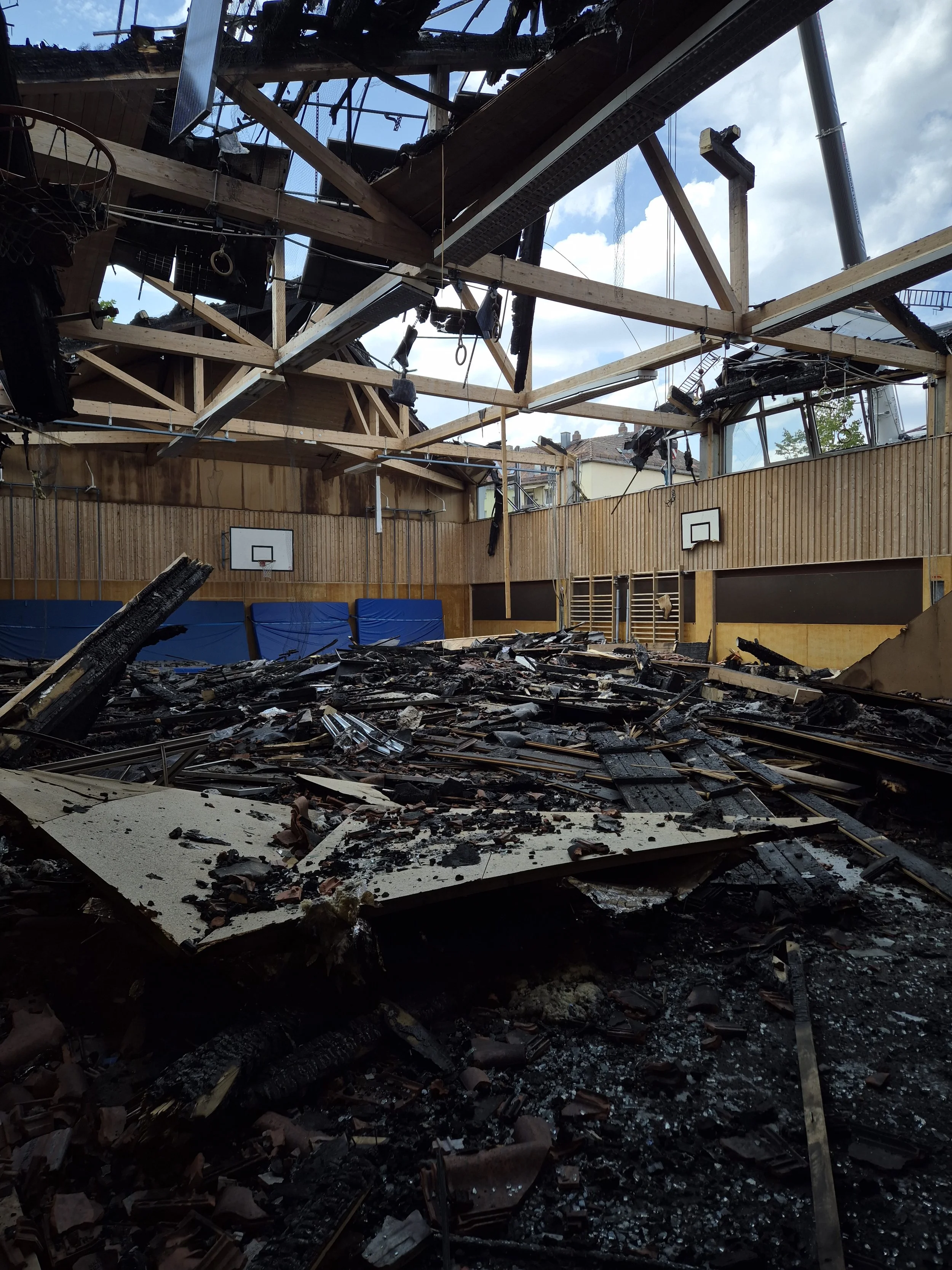 Ausgebrannte Turnhalle mit zerstörtem Dach, verkohlten Trümmern, Basketballkörben und freiliegender Holzkonstruktion