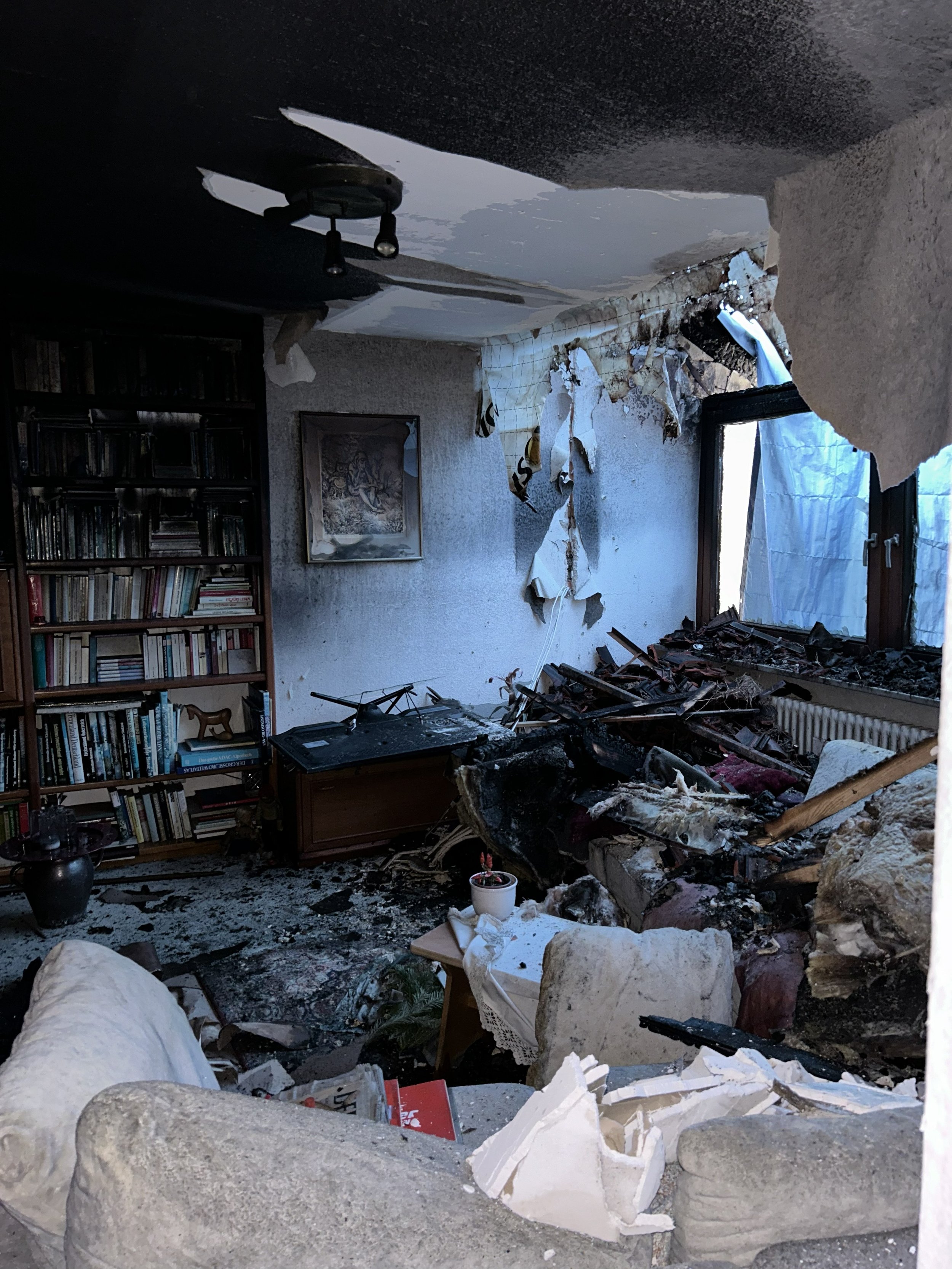 Ausgebranntes Wohnzimmer mit eingestürzter Decke, Brandschutt, beschädigten Möbeln, Bücherregal und freiliegender Satellitenschüssel.
