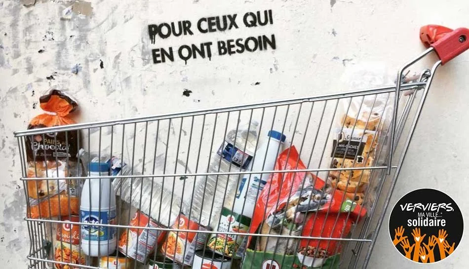 Un caddie de supermarché rempli de nourriture et produits alimentaires, placé contre un mur avec une inscription en français. Logo Solidarité avec des mains levées.
