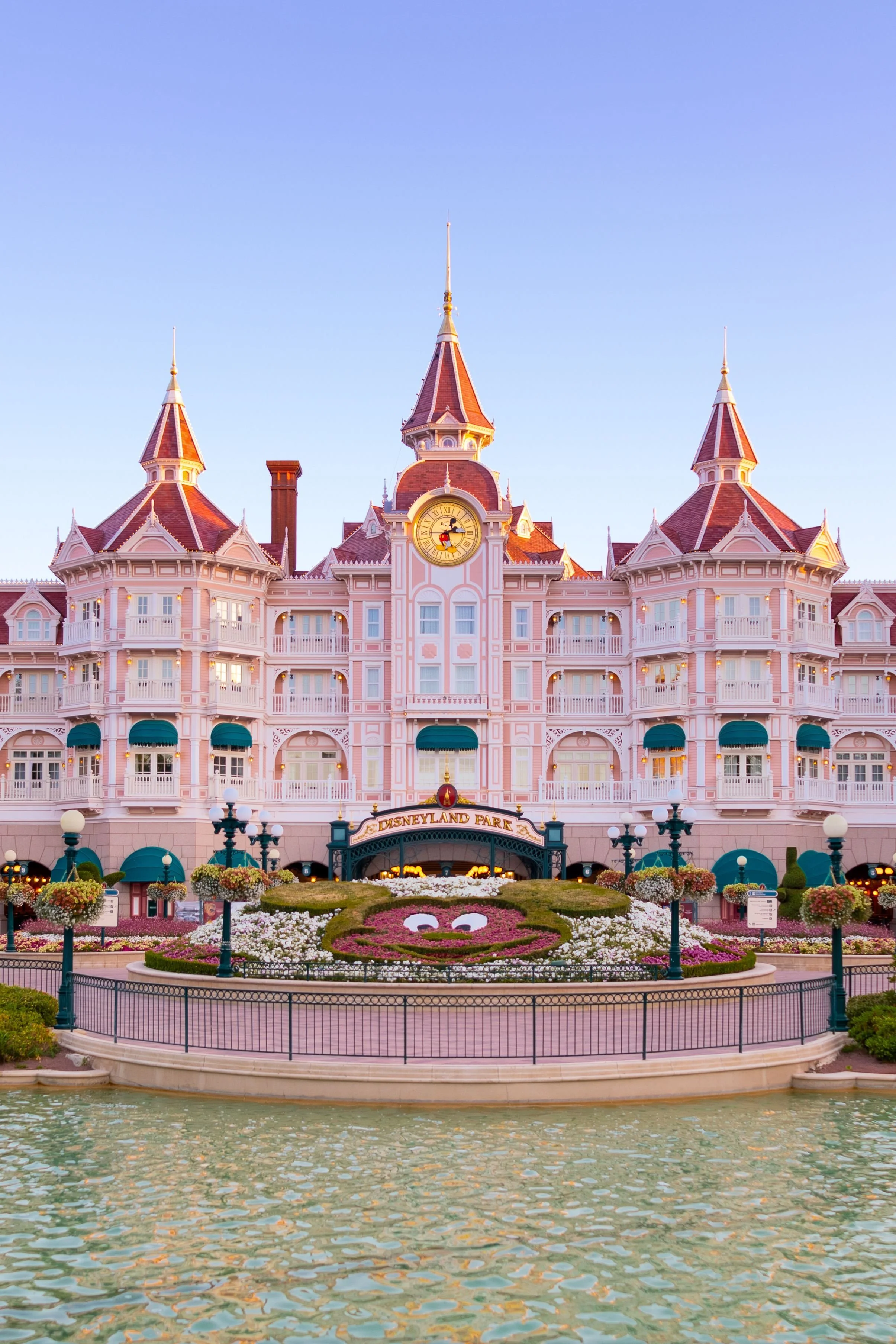 Le château de Disneyland Paris vu depuis le lac avec des fleurs et une fontaine en premier plan, sous un ciel clair.