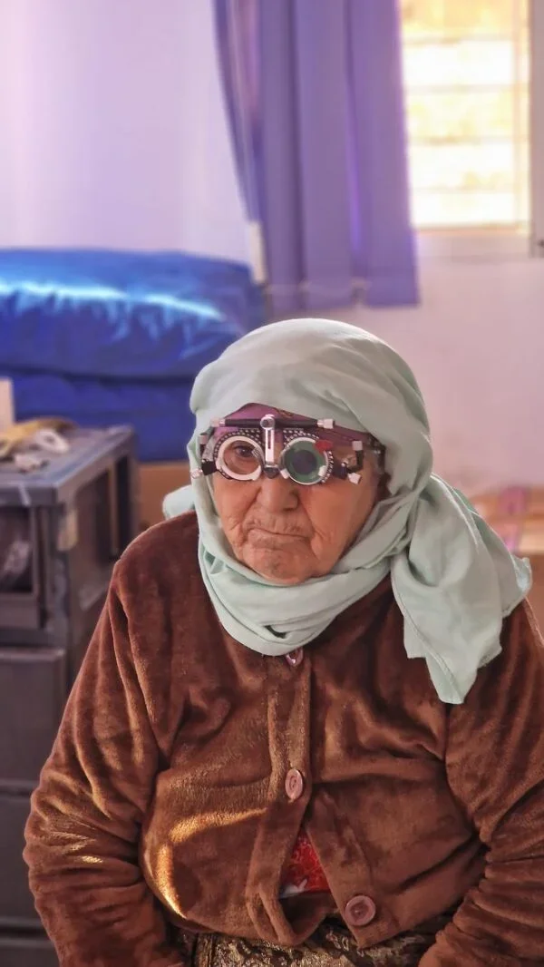 Une femme âgée portant un bonnet gris, des lunettes de presbytie et une robe marron en velours, assise dans une chambre avec un lit en arrière-plan et des rideaux violets.