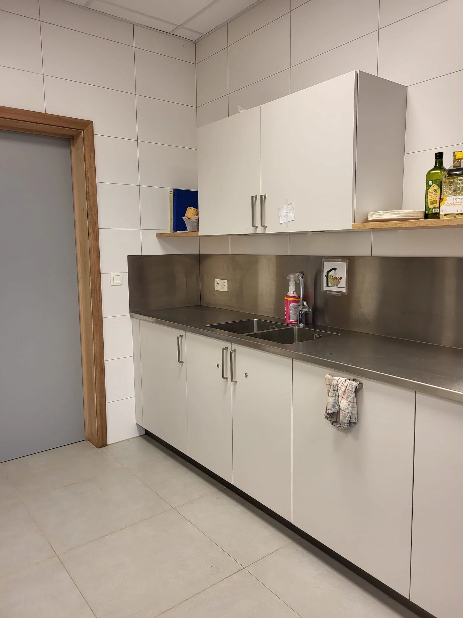 Cuisine moderne avec comptoir en acier inoxydable, armoires blanches, évier double, torchon blanc avec rayures, produits de cuisine sur une étagère, murs en carrelage blanc, sol en carreaux gris clair.