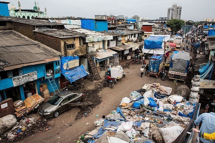 Vue urbaine d'un marché de rue avec des bâtiments en mauvais état, des tentes en tarp, des véhicules, des marchandises empilées, et des personnes marchant dans la rue. En arrière-plan, des immeubles modernes.
