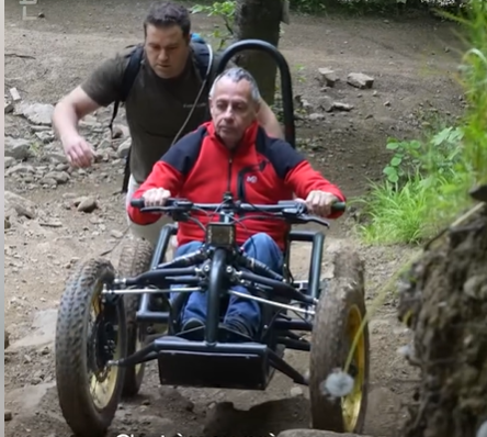 Deux hommes explorent un sentier de forêt, l'un à bord d'un véhicule tout-terrain adapté pour personnes à mobilité réduite, l'autre l'accompagnant en marchant derrière.