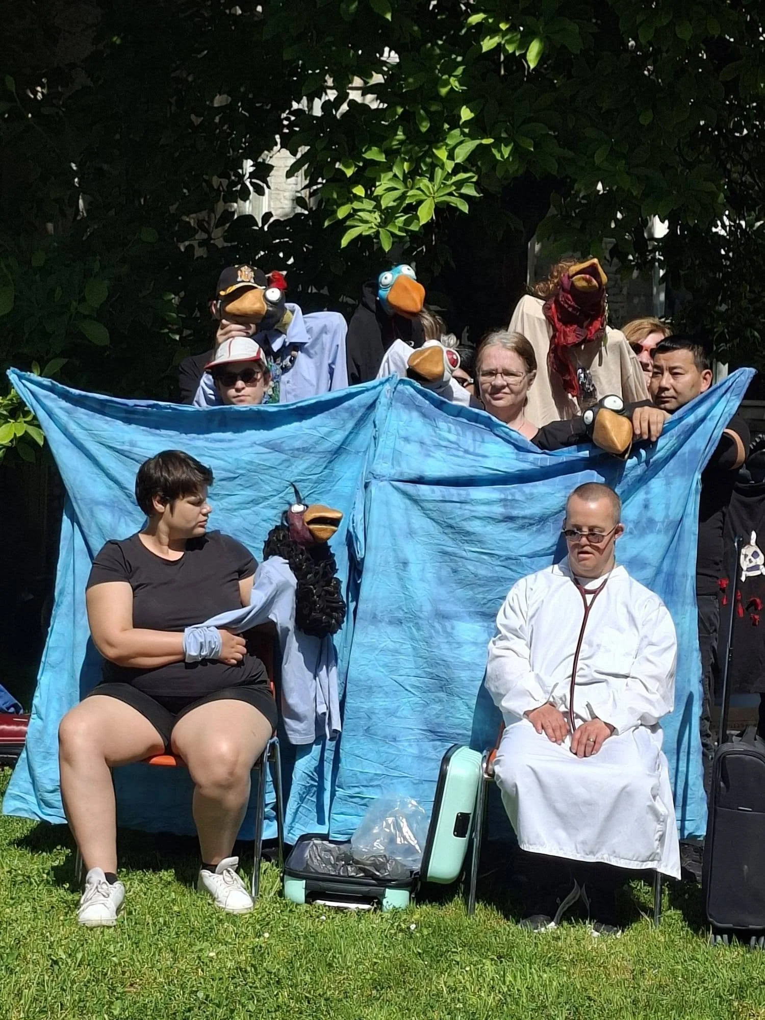 Groupe de personnes portant des masques de silhouettes d'animaux, certains avec des lunettes ou des chapeaux, assises et debout derrière une tente bleue dans un espace extérieur avec de l'herbe et des arbres.