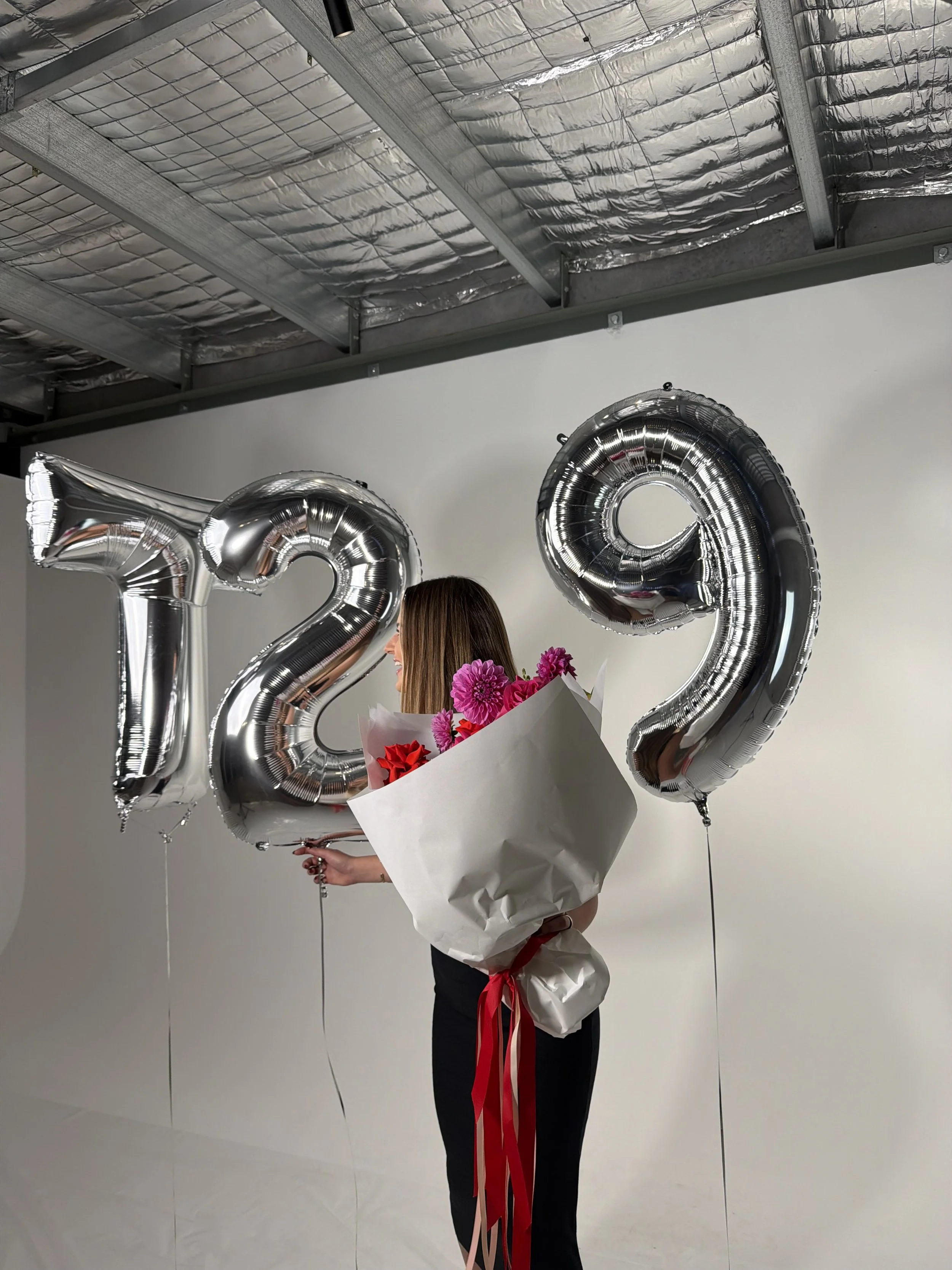 Woman holding a colorful flower bouquet beside large silver number balloons at an indoor celebration