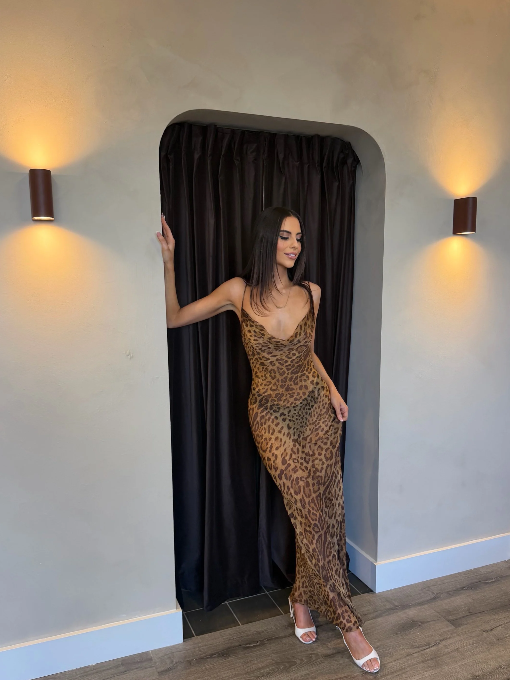Woman posing in a leopard-print dress and white heels in front of black curtains and wall sconces