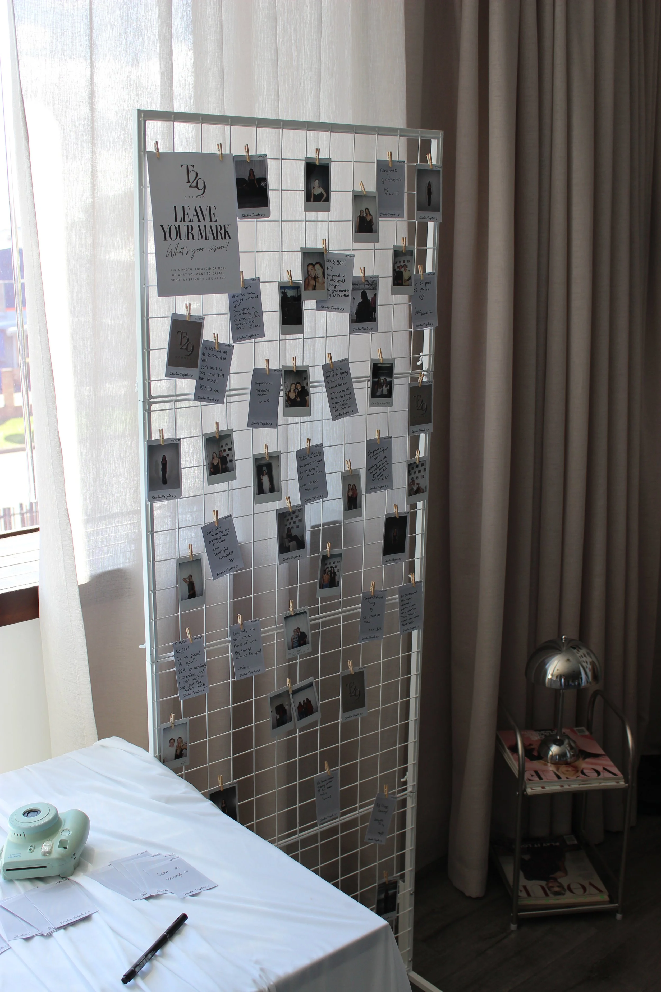 Wire grid display covered with Polaroid photos and handwritten notes beside a table with a Fujifilm Instax camera.