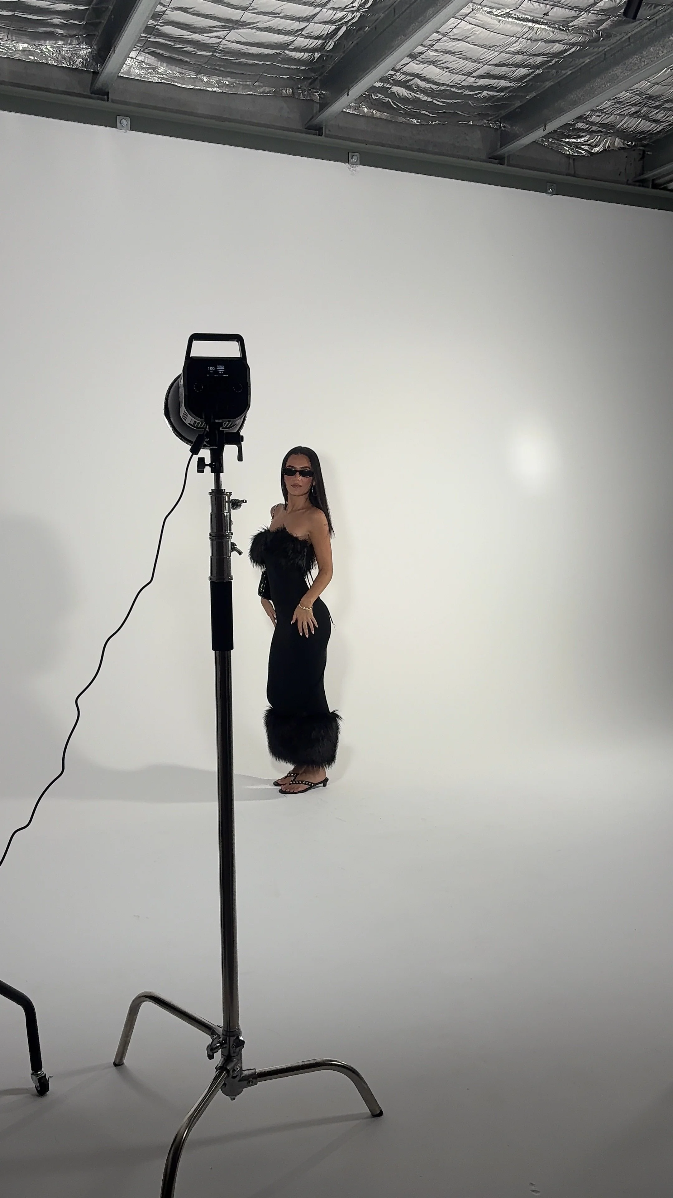 A woman in a black dress with fur trim and sunglasses standing in front of a white backdrop in a photography studio, with a large studio light pointed at her.