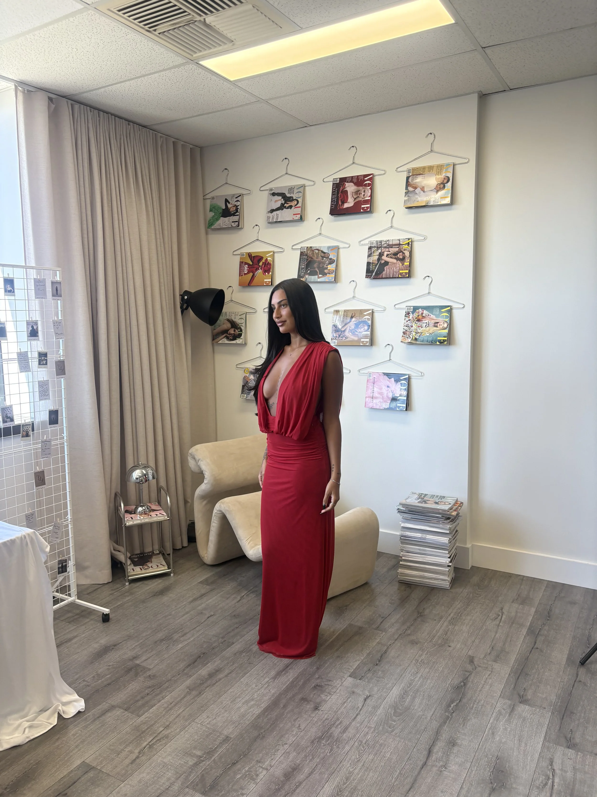 Woman in a red dress standing in a fashion studio beside a wall displaying magazines on wire hangers