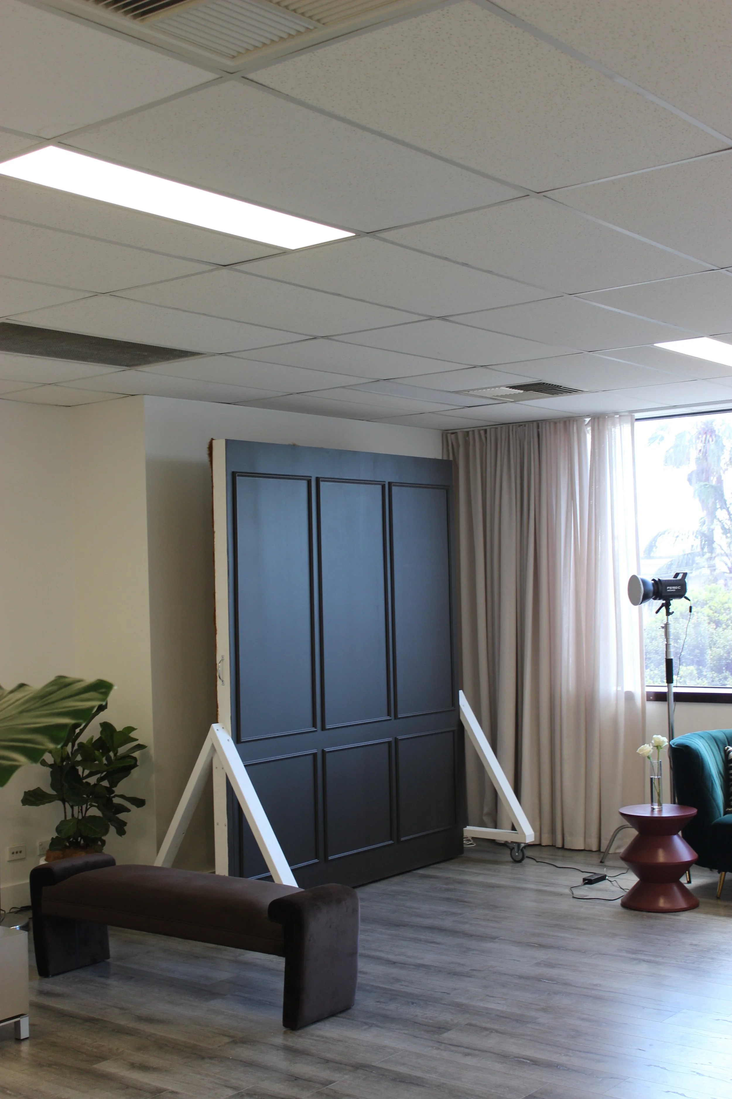 Interior studio with a black paneled backdrop, seating, houseplant, window curtains, and photography equipment.