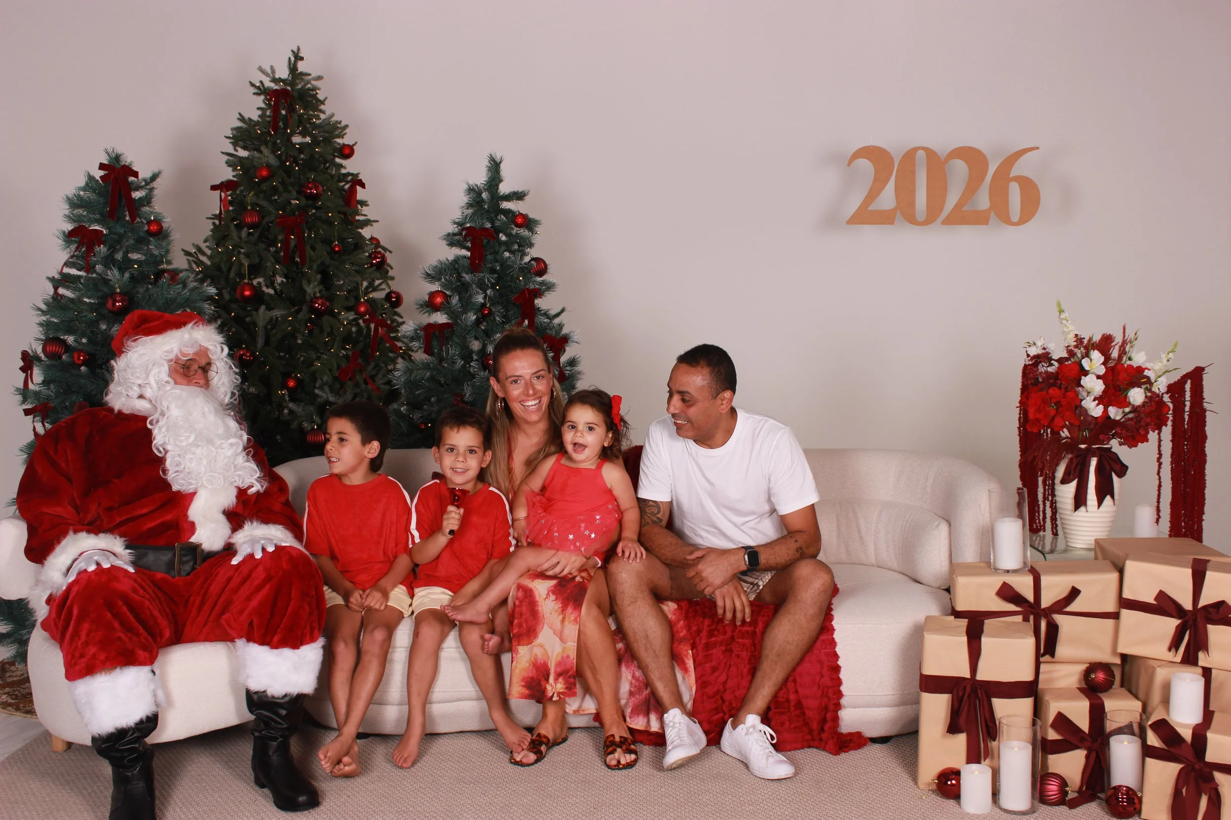 Family posing with Santa Claus beside decorated Christmas trees, wrapped gifts, and a 2026 sign.