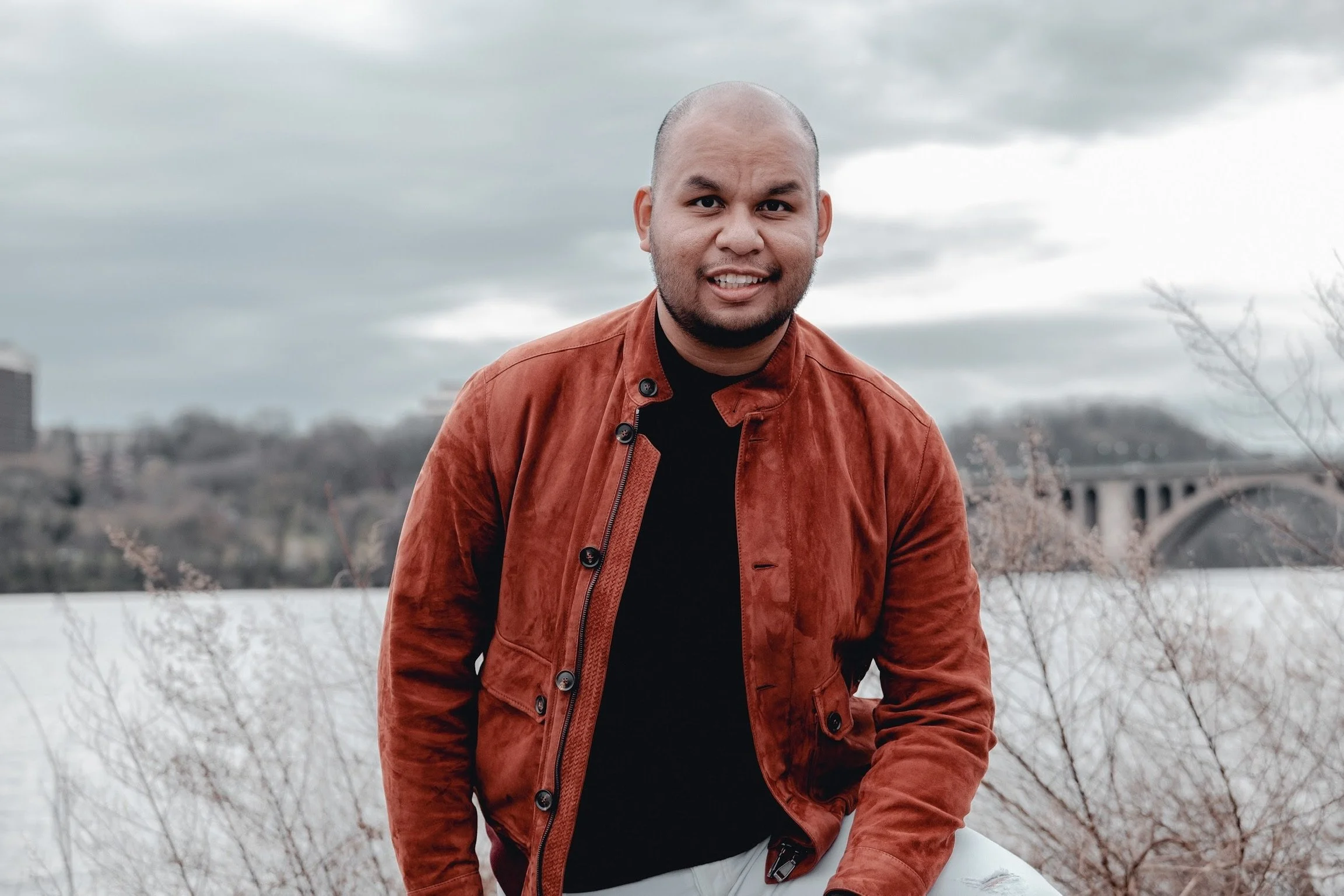 A man with a shaved head and a beard wearing a brown jacket over a black shirt, smiling outdoors with overcast skies and leafless trees in the background.
