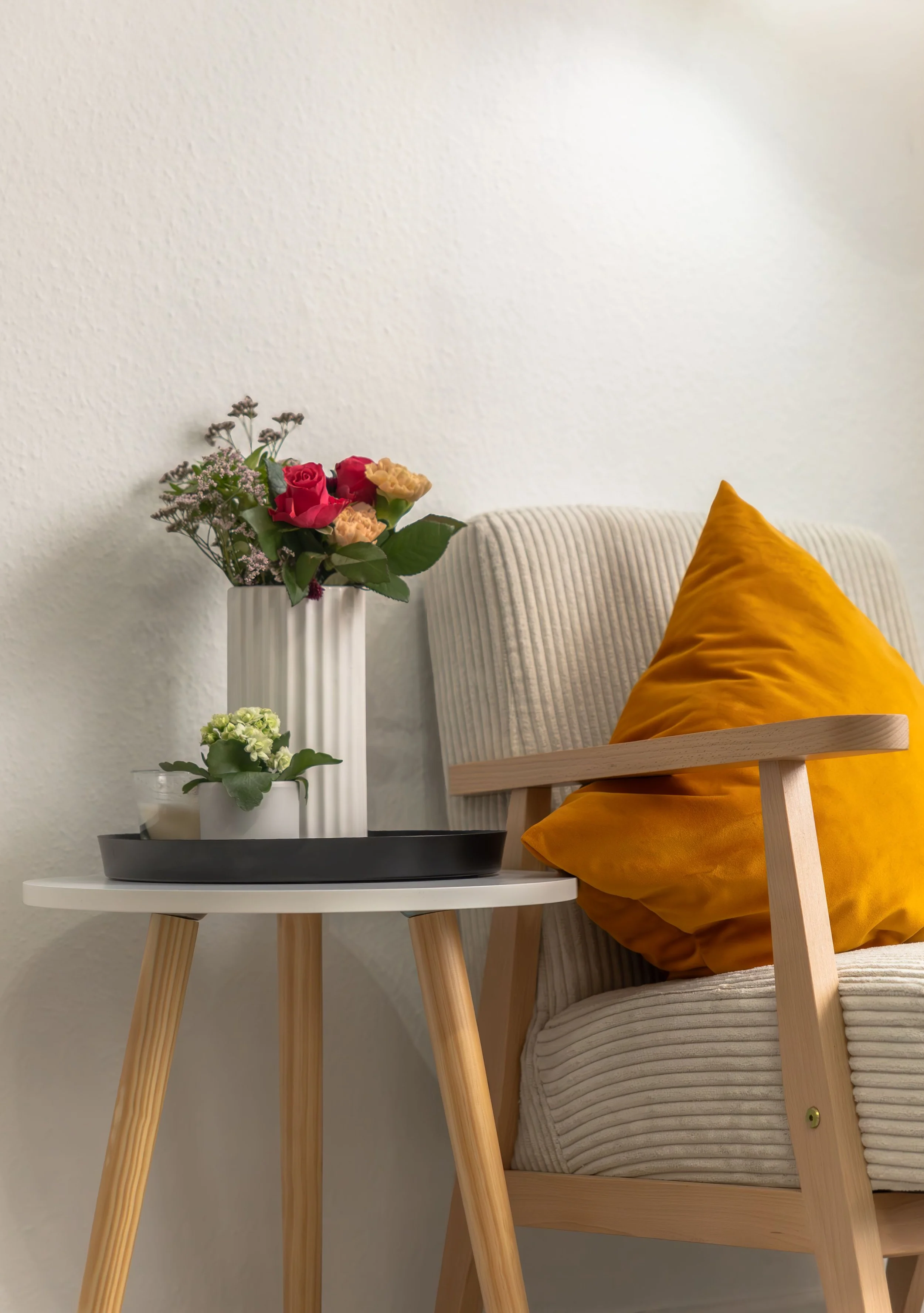 A cozy interior scene with a wooden armchair with a yellow cushion, a small white side table with a black tray, and two potted plants; a larger floral arrangement with pink and peach flowers in a white striped vase, and a smaller green plant in a white pot, against a plain white wall.