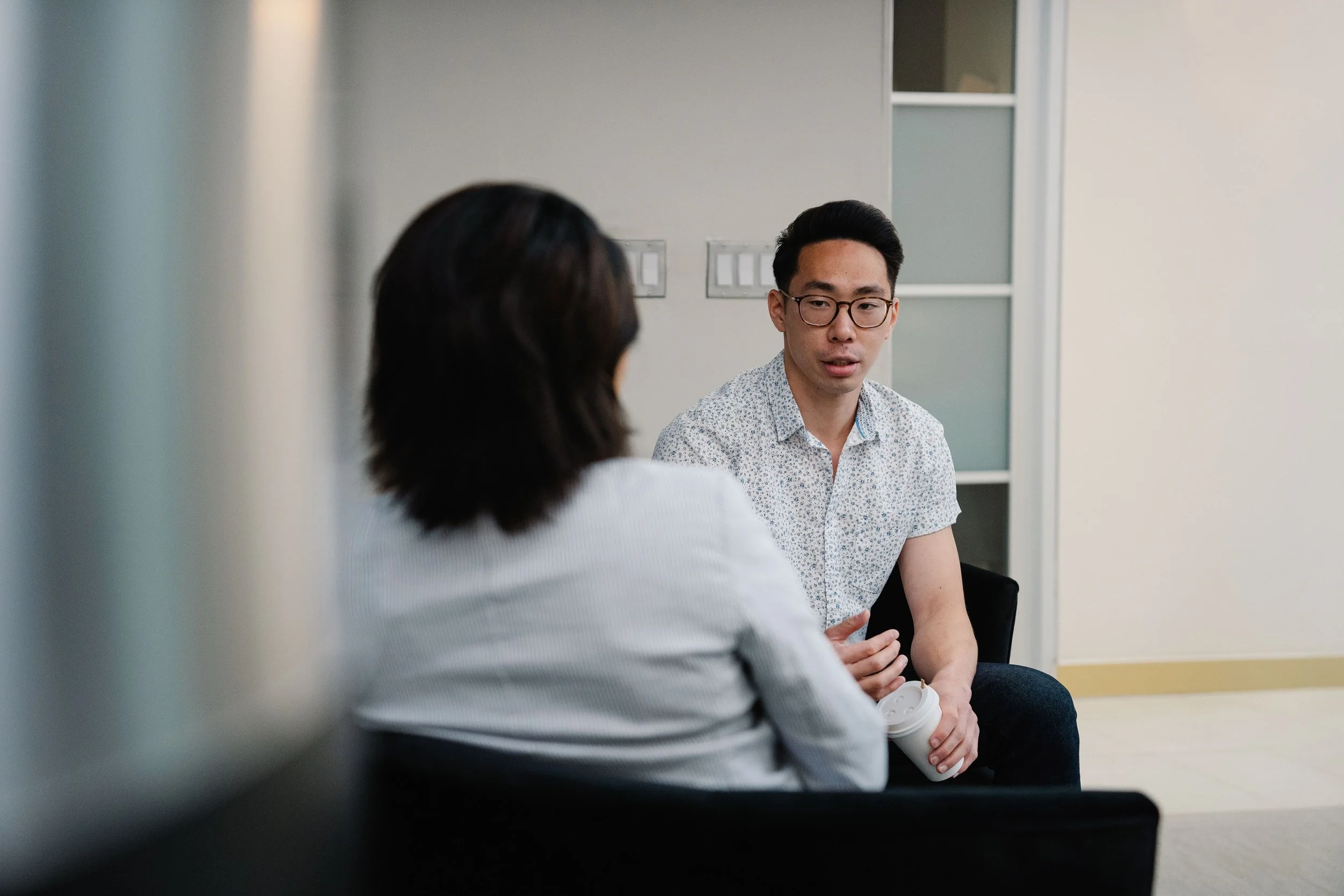 Two people, a man with glasses wearing a patterned shirt and a woman with dark hair, are seated and having a conversation in a modern room. The man is holding a coffee cup, and the woman has her back to the camera.