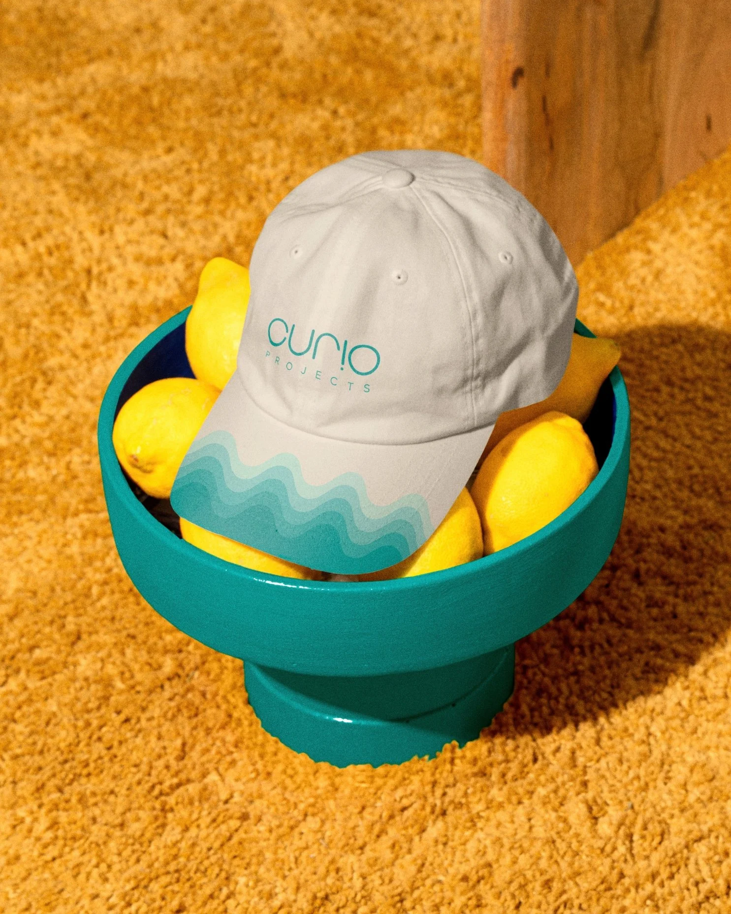 A blue bowl filled with lemons with a white cap on top that has the text 'Curio Projects' written on it, placed on a carpeted floor.