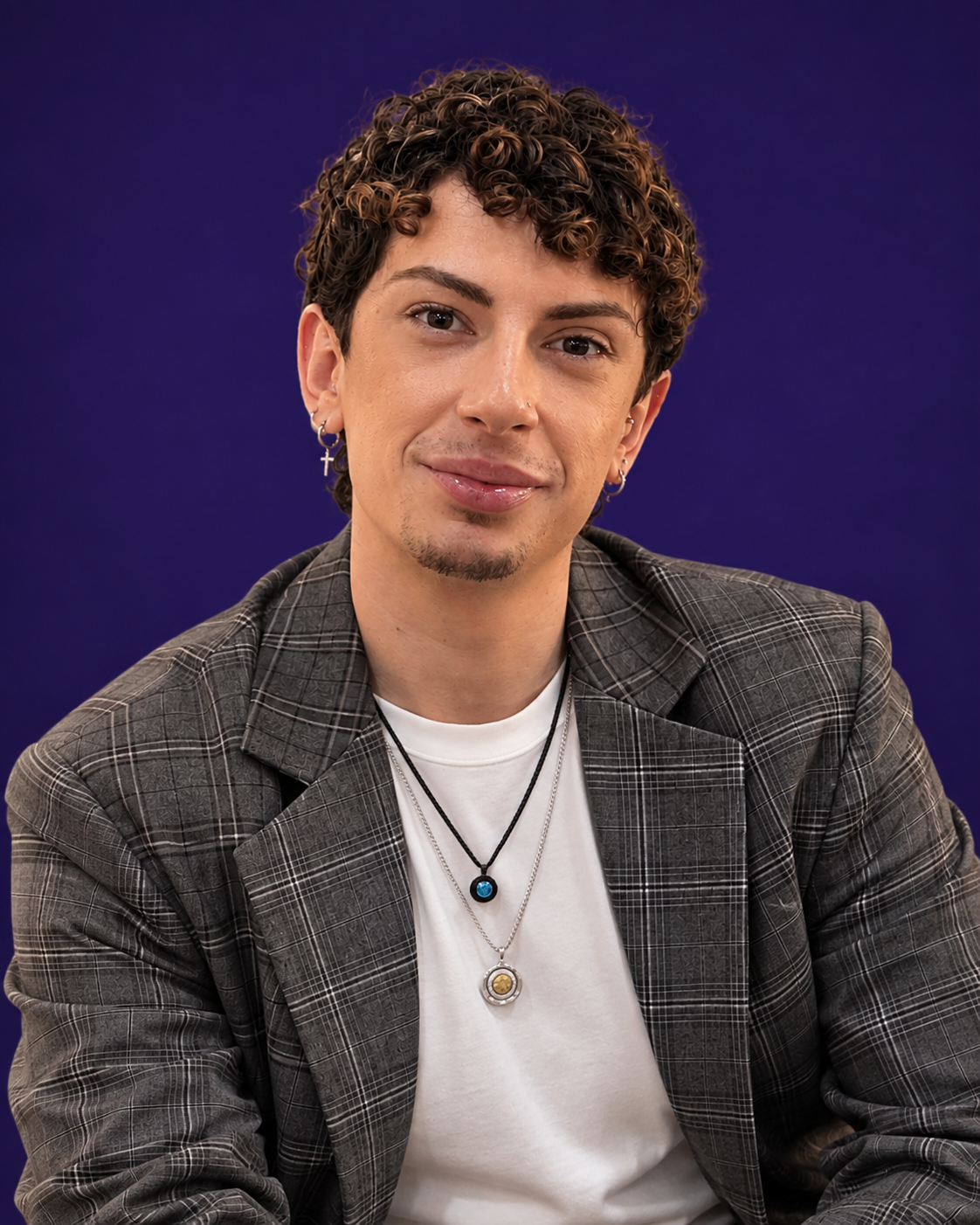 A young man with short curly hair, wearing earrings, necklaces, a white t-shirt, and a plaid jacket, smiling in front of a solid dark blue background.