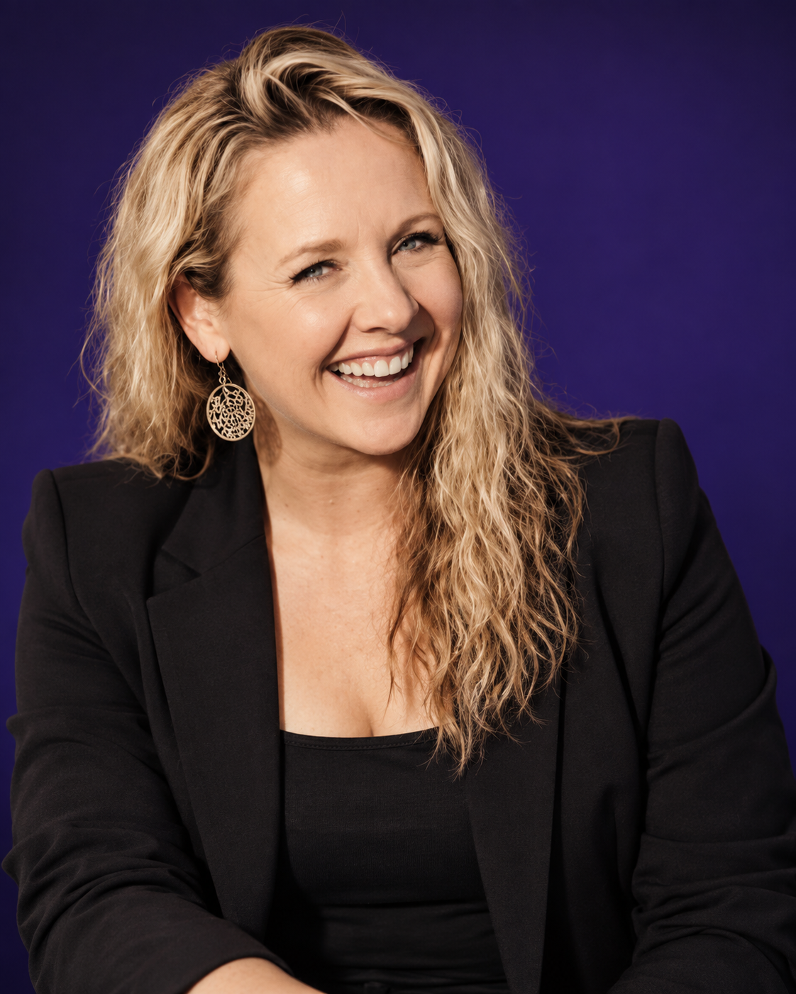 Smiling woman with curly blonde hair wearing a black blazer and large circular earring against a blue background.