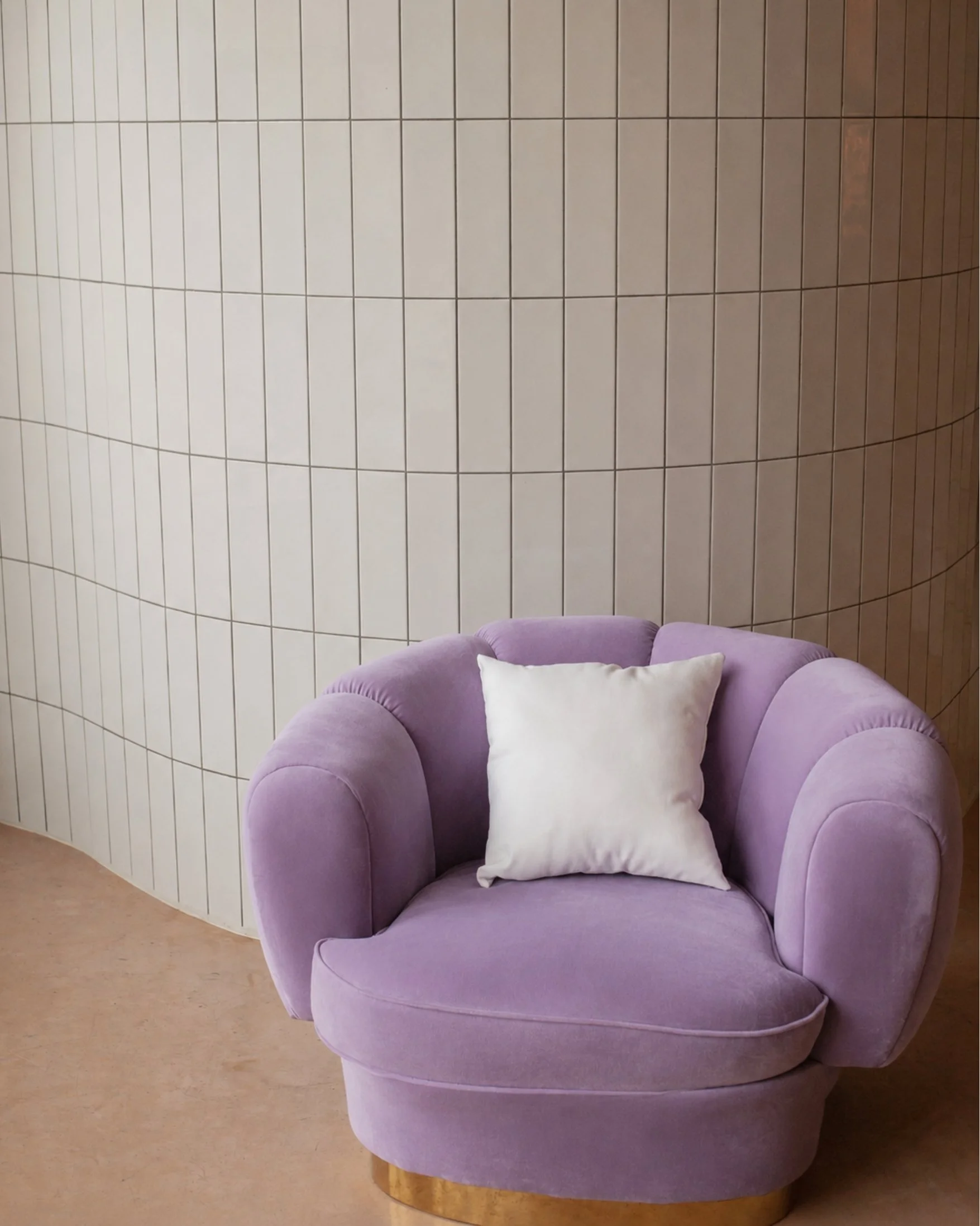 A purple upholstered armchair with a white pillow on it, placed in front of a curved, beige tiled wall.