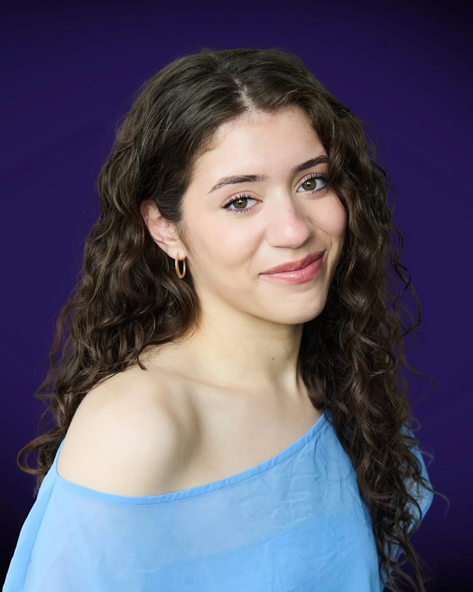 A woman with wavy brown hair, wearing a light blue off-the-shoulder top and a gold hoop earring, looking at the camera against a dark purple background.