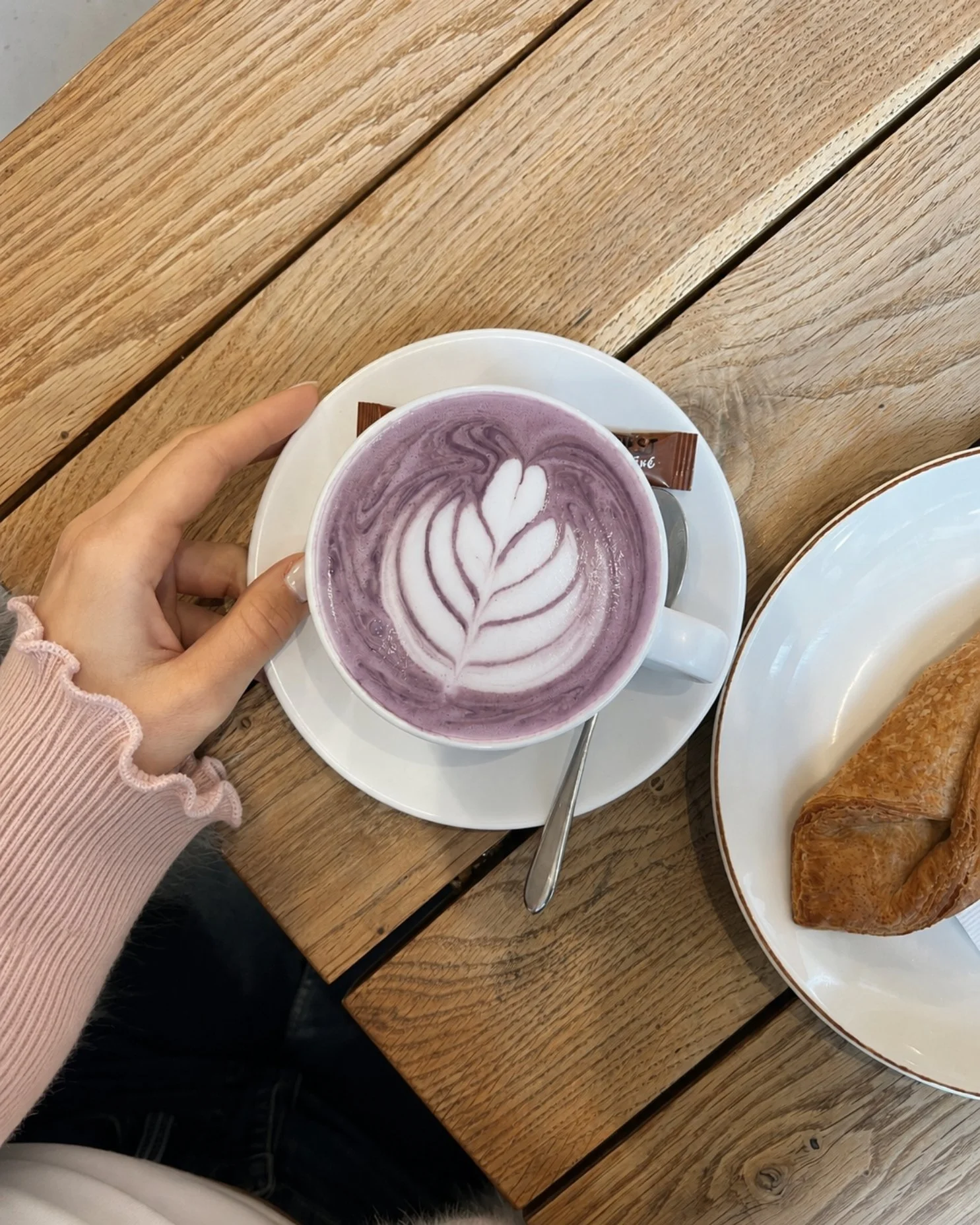 A person holding a cup of purple latte with latte art on top, on a wooden table with a plate of croissants.