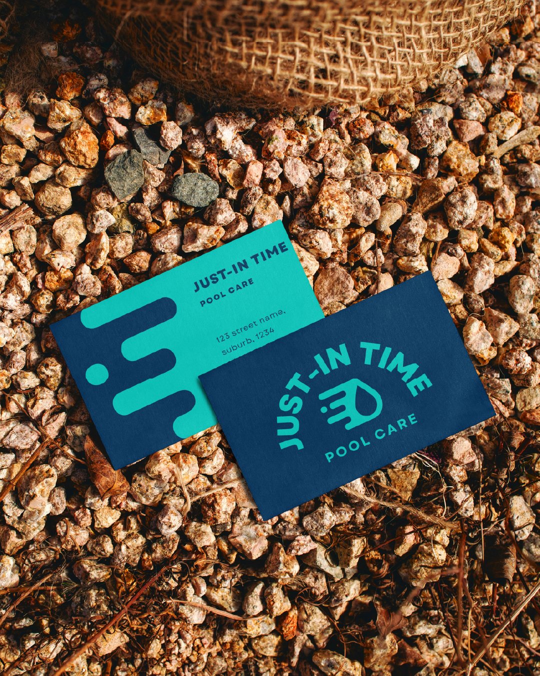 Two business cards for 'Just-In Time Pool Care' on rocky ground, one blue with logo and one turquoise with contact information.
