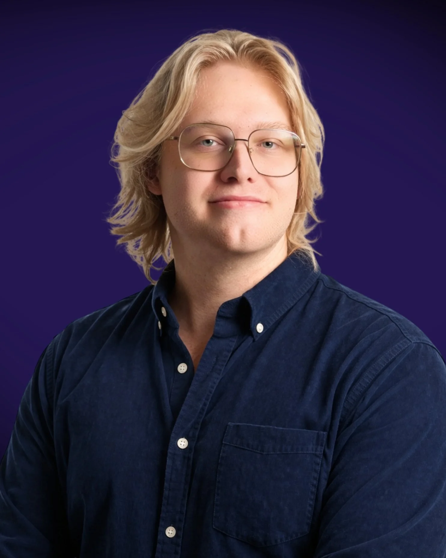 A young man with shoulder-length blond hair wearing glasses and a dark blue button-up shirt, posing against a dark purple background.