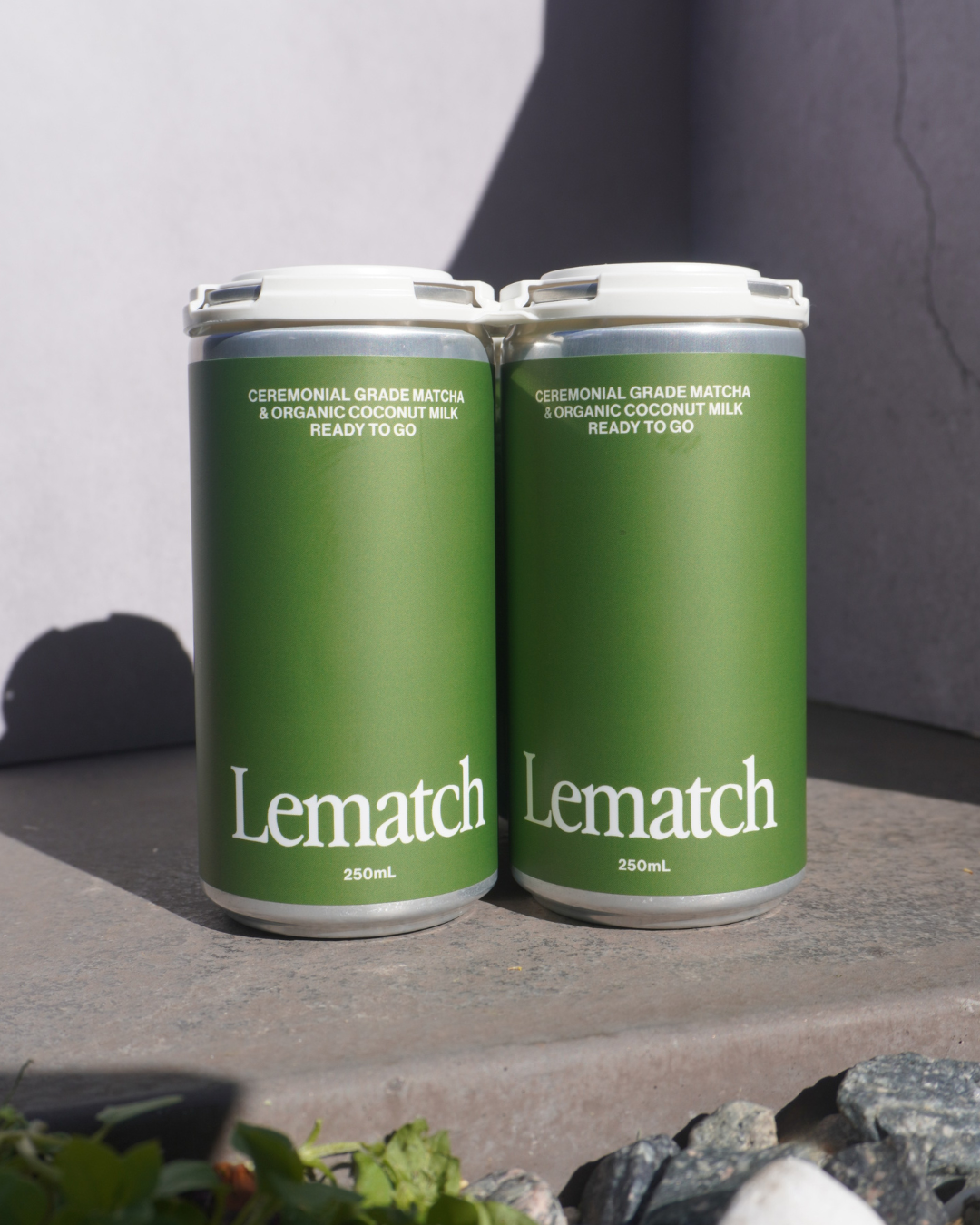 Two cans of Lematch beverage with green labels on concrete surface, sunlight, and shadows.