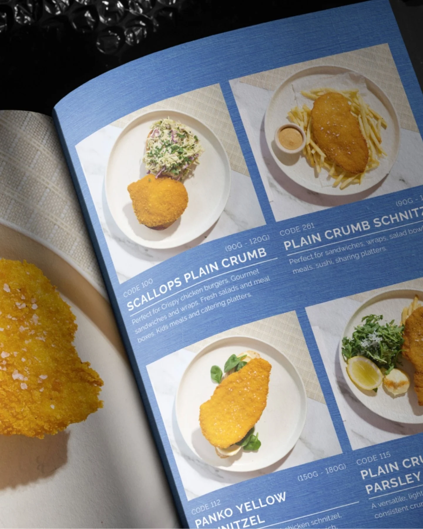 Close-up of a restaurant menu page showing two dishes: on the left, a breaded fried item with a side salad; on the right, a breaded fried item with French fries, a small cup of sauce, and a lemon wedge, with partial view of a fried item on a plate in