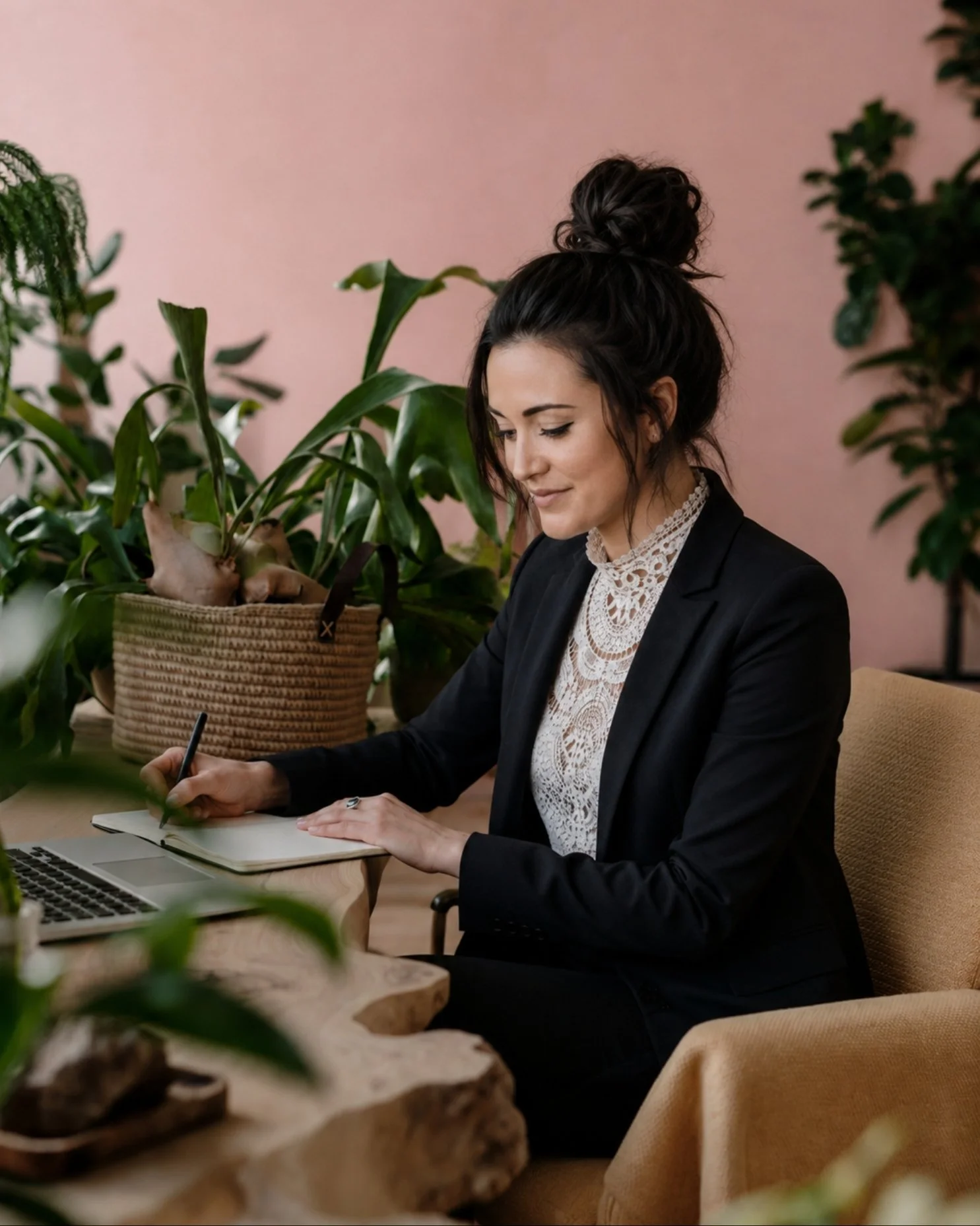 A woman in business attire sitting at a wooden table with green plants around, working on a laptop and writing in a notebook, against a pink wall background.