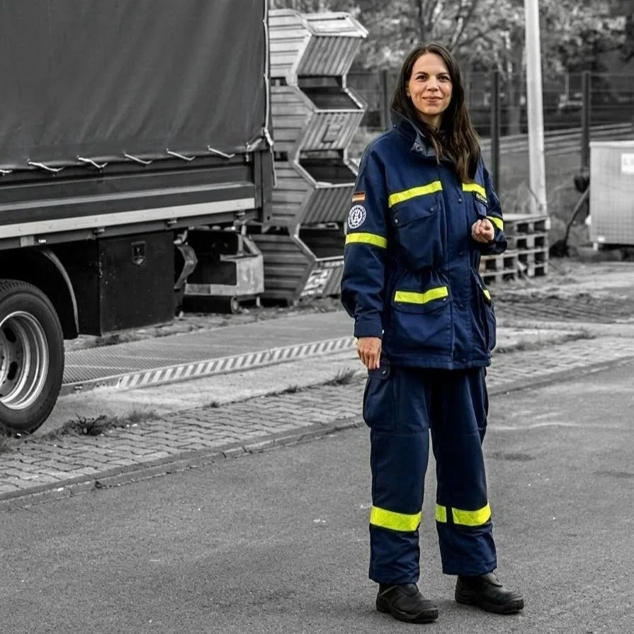 Eine Frau in einer dunkelblauen Einsatzuniform mit reflektierenden Streifen steht auf der Straße, im Hintergrund sind Lagerregale und ein Transit-LKW zu sehen.