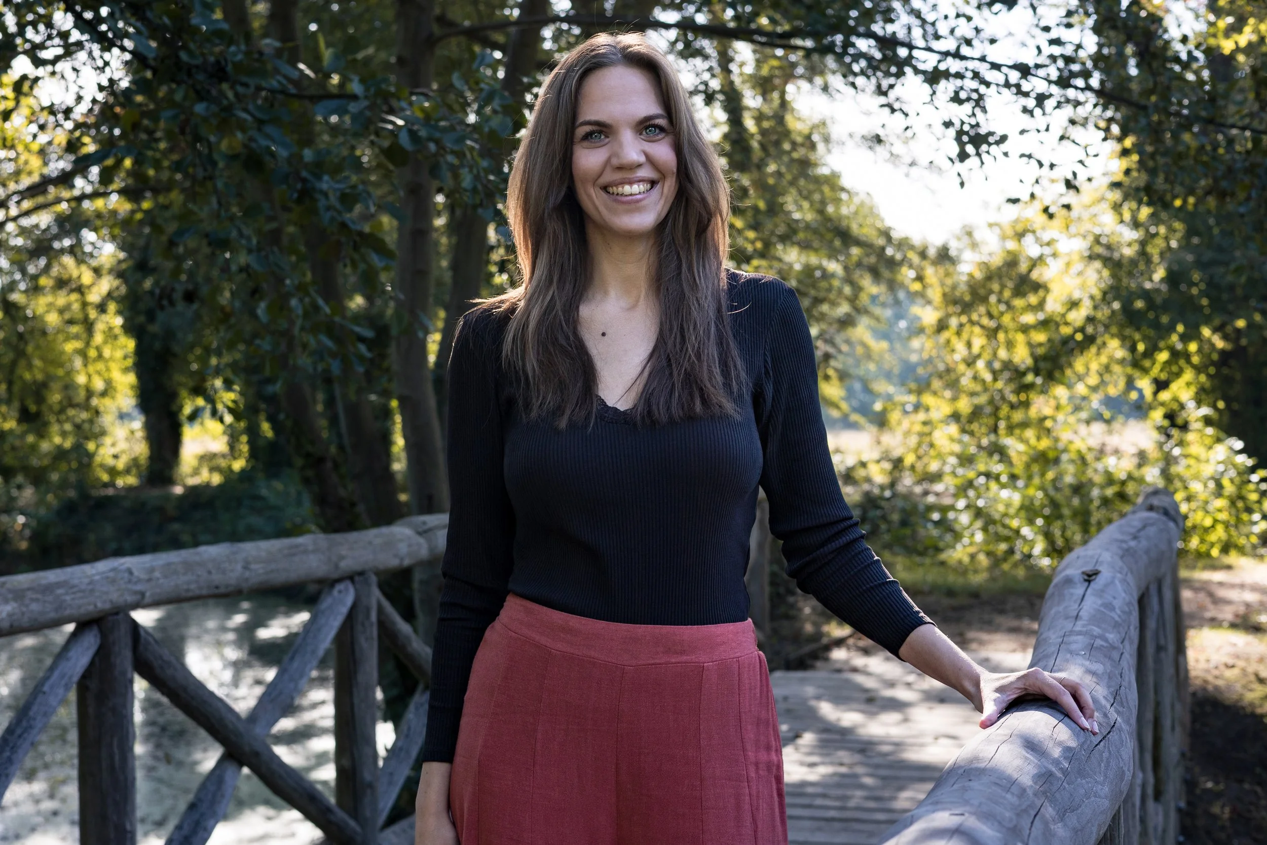 Eine junge Frau mit braunen, langen Haaren, blauen Augen und einem Lächeln steht an einem Waldweg neben einem Holzzaun, im Hintergrund grüne Bäume, sonniges Wetter.