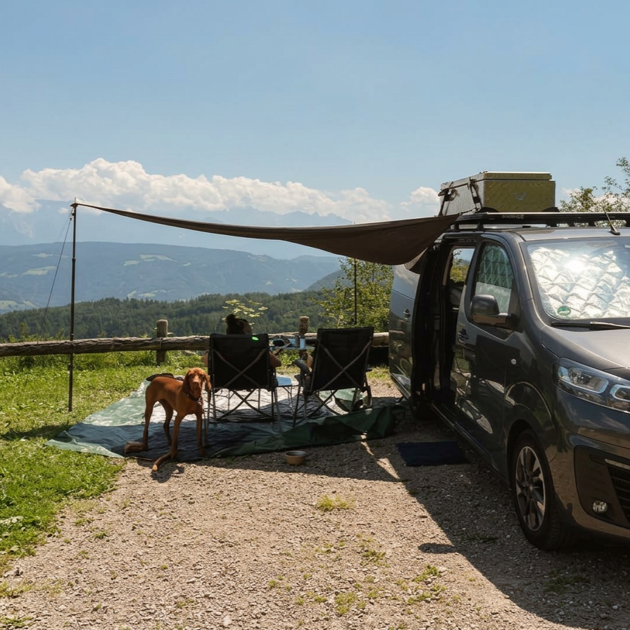 Zwei Menschen sitzen in Campingstühlen vor einem Campingwagen in der Natur, mit Blick auf Berge und Himmel. Ein Hund sitzt auf einer Matte neben den Stühlen.