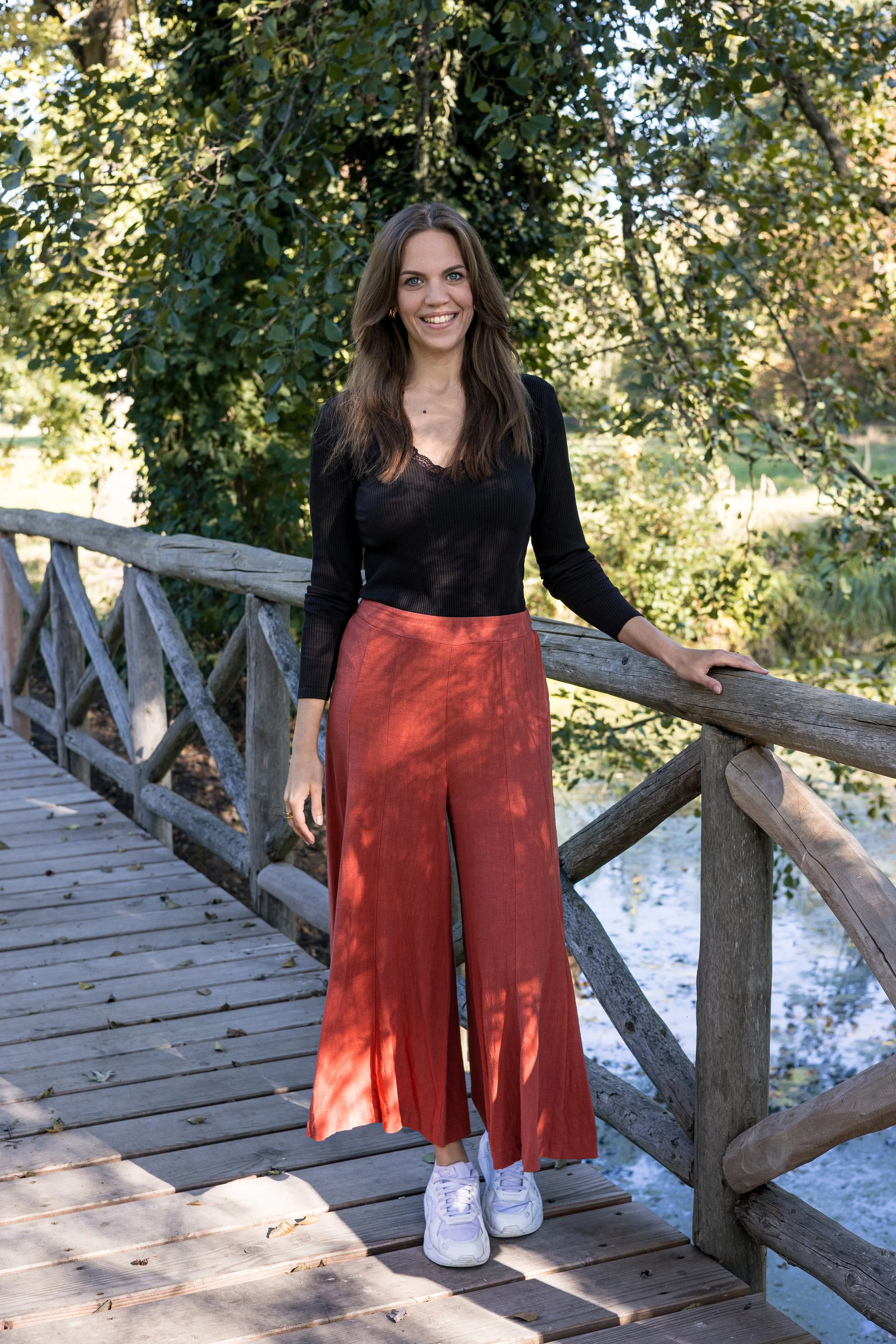 Junge Frau mit braunen Haaren, schwarz-weißes Oberteil, roten weiten Hosen und weißen Sneakers steht auf einer Holzbrücke neben einem Wasserlauf, im Hintergrund Bäume und Natur bei Sonnenlicht.