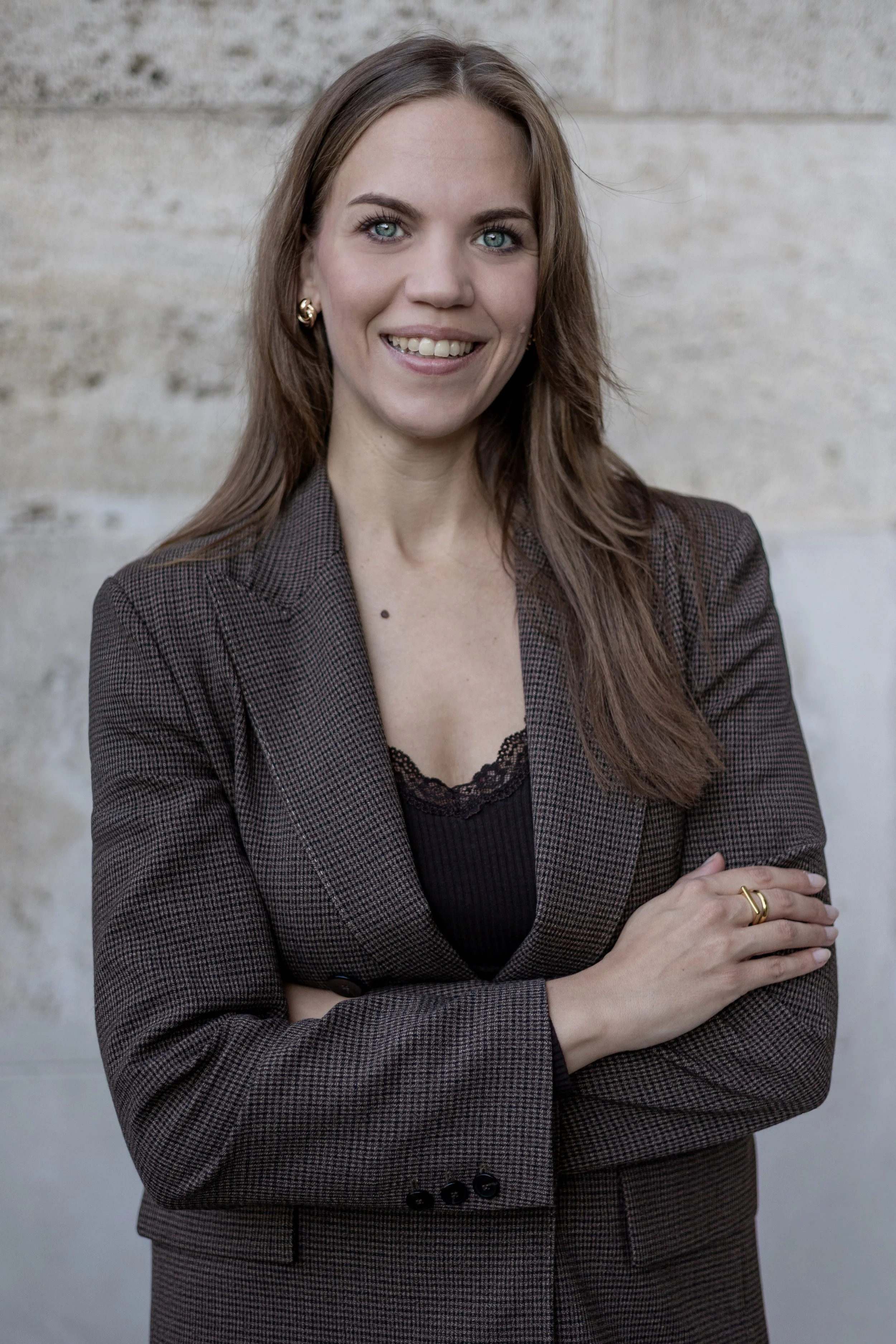 Eine lächelnde Frau mit blauen Augen, braunen Haaren und goldenen Ohrringen, trägt einen blass braunen karierten Blazer und ein schwarzes Oberteil, steht vor einer Betonmauer.