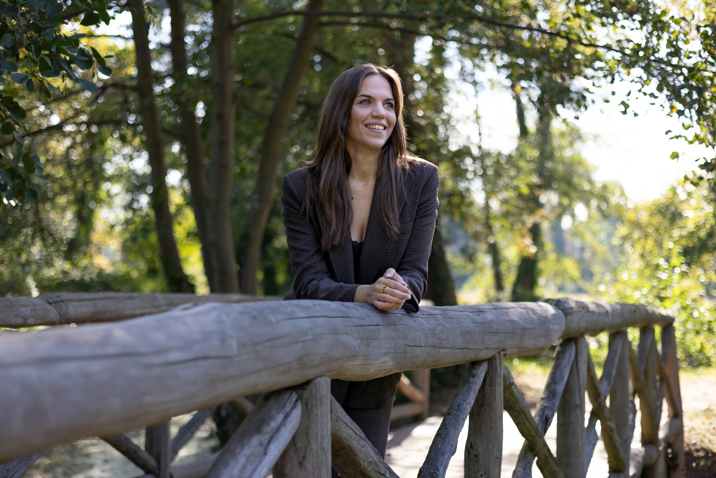 Eine junge Frau mit dunklem, langem Haar, die lächelnd in einem Park auf einer Brücke aus Holz und Holzpfählen steht, umgeben von Bäumen mit grünem Laub und Sonnenlicht, das durch die Blätter scheint.