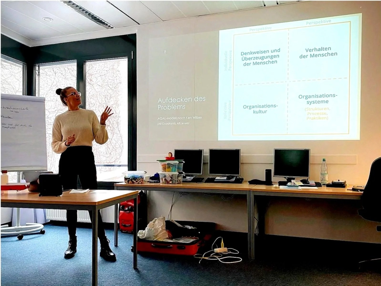 Eine Frau präsentiert eine Folie in einem Seminarraum. Im Raum stehen Tische, auf denen Computer, Büromaterialien und persönliche Gegenstände liegen. Die Präsentation zeigt ein Diagramm zu Organisationsthemen.